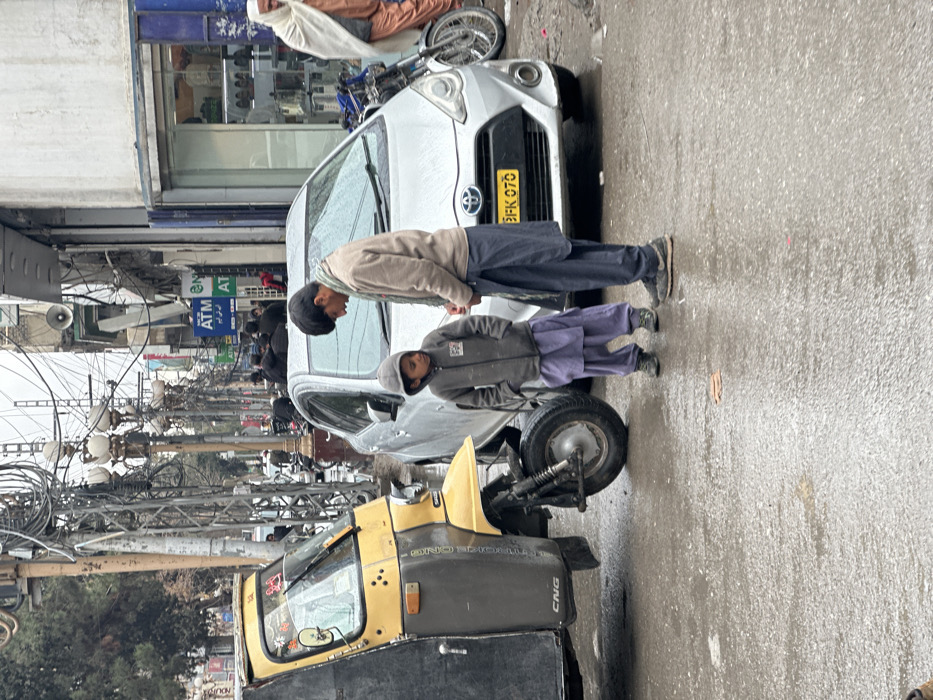 The image captures an urban street scene in Quetta City Tehsil, Pakistan, on a damp, overcast day, evidenced by the wet asphalt road. In the foreground, two individuals, an adult male and a young child, stand facing each other on the wet pavement. The adult male wears a beige jacket over a patterned scarf and dark blue shalwar kameez, looking down at the child. The child, approximately 5-8 years old, wears a grey hooded jacket and light purple shalwar, with hands in pockets, looking up at the adult.
To the left of the individuals, a yellow and black three-wheeled auto-rickshaw with "CNG" visible is parked. Immediately behind them, a silver Toyota Aqua hatchback, bearing license plate "3FK 070," is parked. On the far right, partially visible, is an older male figure in a white shawl and orange shalwar kameez, with a motorcycle parked nearby. The background reveals a bustling street lined with commercial buildings, identifiable by storefronts and signage, including an "NCB ATM." The scene is characterized by numerous utility poles with dense overhead wiring and ornate streetlights extending down the thoroughfare.