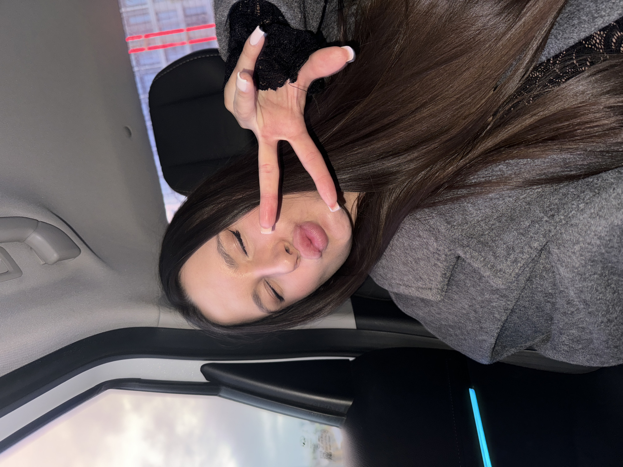 The image depicts a person with long, dark brown hair lying inside a car. The person is wearing a gray coat and a black lace sleeve. The person is making a peace sign with their right hand near their face, while their eyes are closed and their lips are slightly pursed. The background visible through the car windows appears to be urban, possibly indicating a city setting. There is no evidence the image was taken in Moscow, Russia.