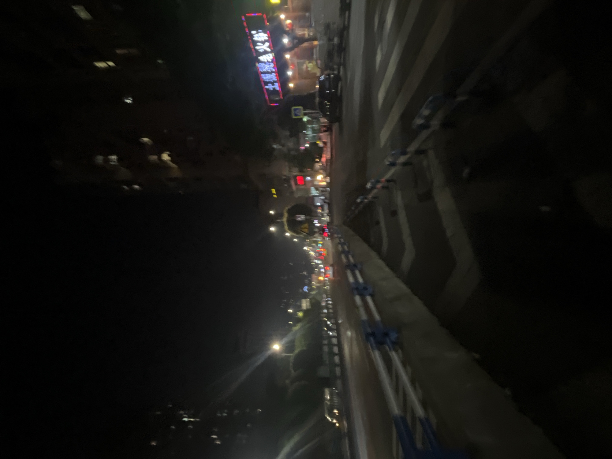 The image captures a nighttime view of a street in Shapingba District, China. The scene is dominated by streetlights and the illuminated signs of businesses. In the foreground, a guardrail with blue and white markings lines the side of the road. A pedestrian crossing is visible on the right side of the image. Several vehicles, including cars and what appears to be a bus, are on the road. On the left, a hillside is visible with scattered lights, suggesting buildings or homes. In the background, a tall building with illuminated windows stands against the dark sky. A prominent neon sign in Chinese characters is visible above a storefront. The overall atmosphere is that of a busy urban street at night.