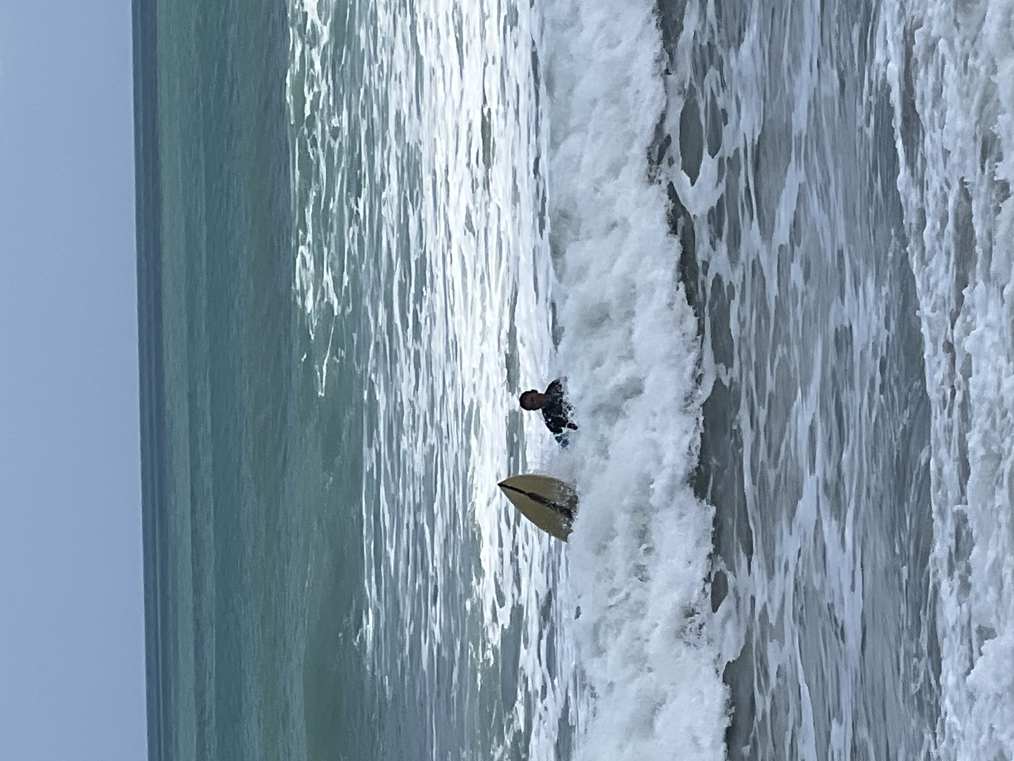 A surfer in a black wetsuit is in the ocean, partially submerged in a wave with a yellow surfboard. The surfer is facing forward, looking towards the viewer. The surfboard is upright in the water, with the nose pointing towards the sky. The ocean is choppy with white foam from the waves. The background shows the ocean stretching to the horizon, with a clear sky above. This scene is located in the city of Kuta, Indonesia.