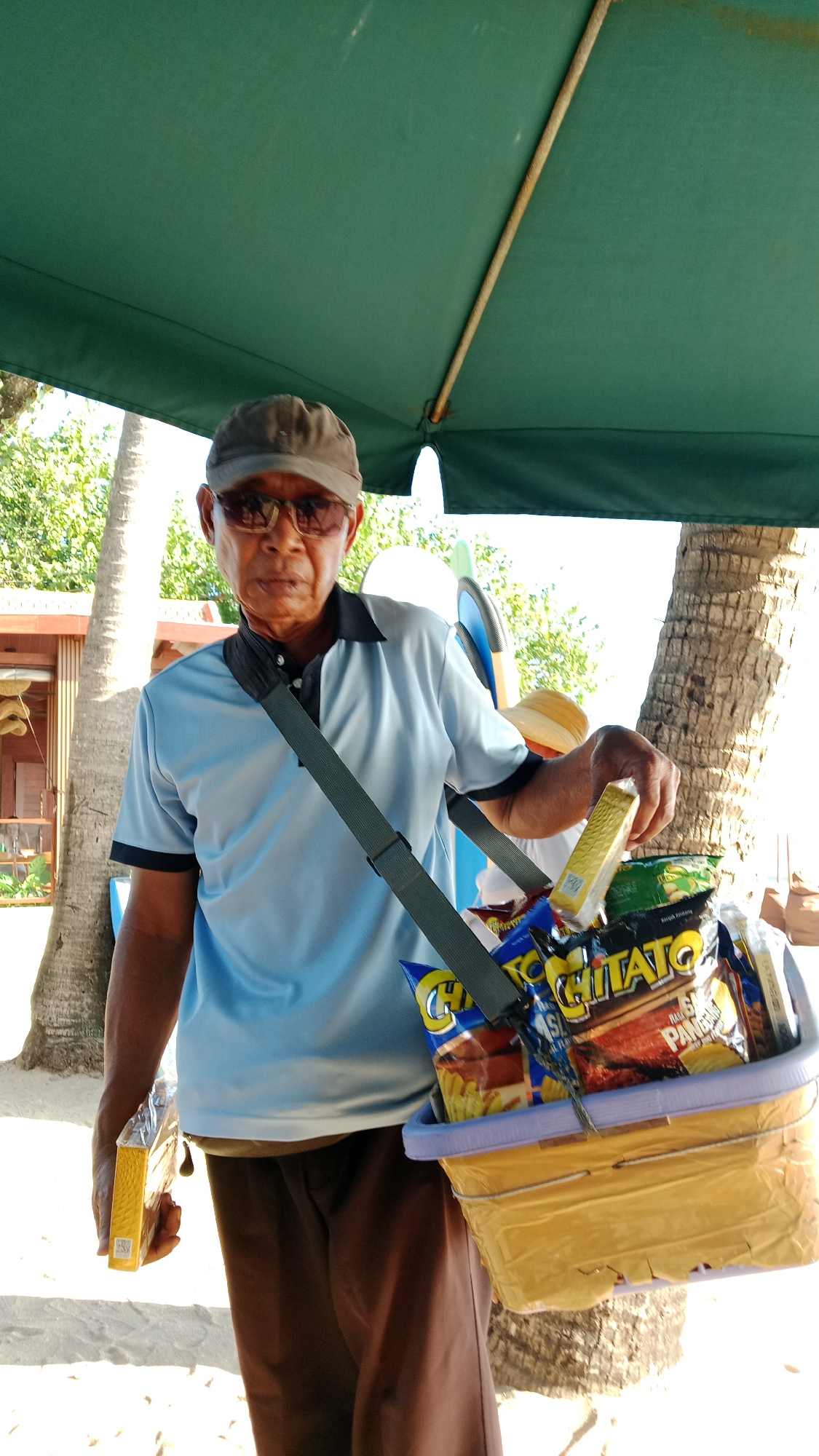 An elderly East Asian man wearing a light blue polo shirt, brown pants, a cap, and sunglasses stands outdoors under a green awning. He is holding a yellow box in his left hand and a yellow basket filled with snacks on his right side. The basket is slung over his shoulder by a dark strap. The man appears to be selling snacks on a beach or in a tropical area, with palm trees and a sandy ground visible in the background. The overall lighting suggests it is daytime.