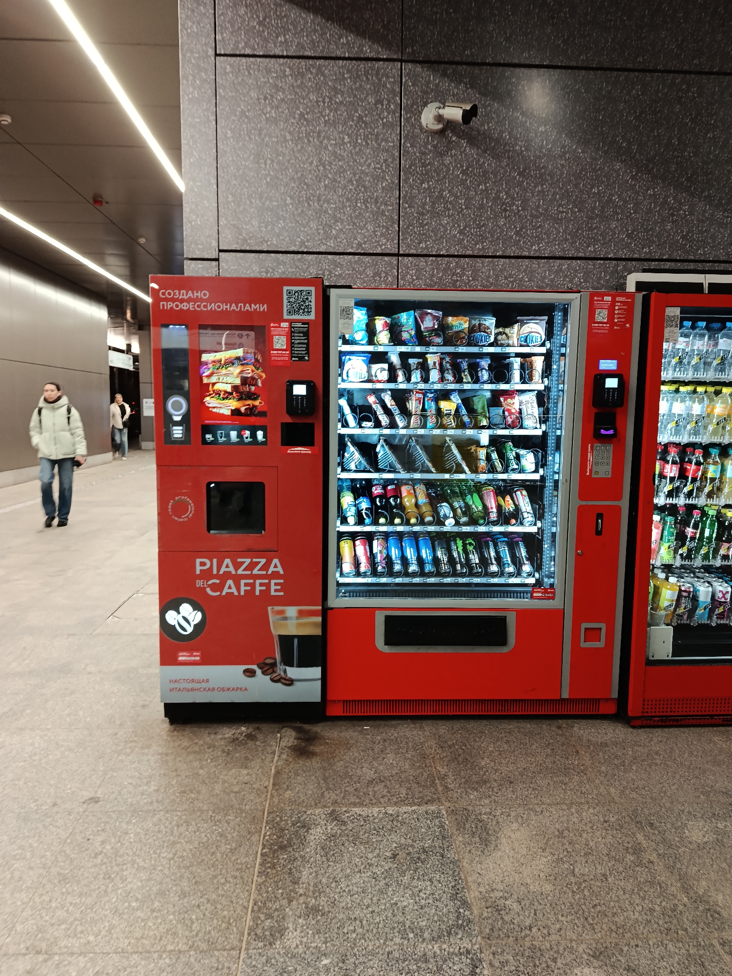 The image shows a public area, possibly a subway station in Moscow, Russia. The environment includes light-colored tile flooring and gray wall panels. Two people are walking away in the background. In the foreground, there are three vending machines. The first is a red coffee vending machine that says "PIAZZA DEL CAFFE." The second vending machine is also red and stocked with snacks and drinks. The third vending machine is red and appears to have bottled drinks. Above the vending machines on the wall, there is a security camera.