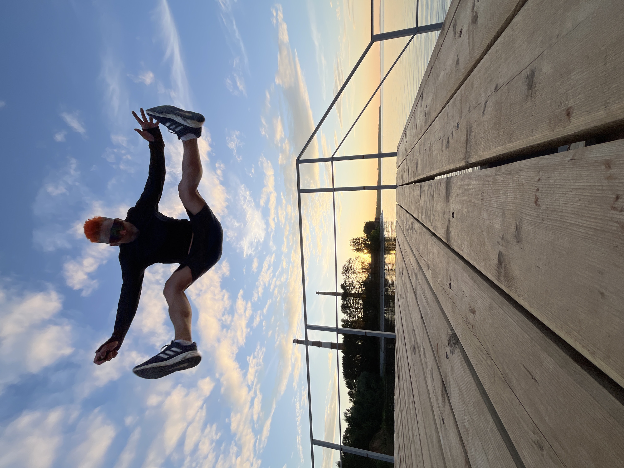 A low-angle photograph captures a male subject performing a straddle jump over a wooden pier near Chaikovsky, Russia, during the golden hour. The subject is airborne, centered against a vibrant sky with scattered cumulus clouds illuminated by warm, golden light near the horizon. He has distinctive bright orange, spiky hair and wears dark sunglasses. Dressed in a black long-sleeved athletic top, black shorts, and black Adidas sneakers with white soles, his arms are extended outwards with open hands, and his legs are spread wide in a horizontal split.
The foreground consists of light-colored wooden planks of a pier, showing grain and texture, extending from the bottom of the frame towards the distant horizon. Metal handrails, composed of vertical posts and horizontal bars, ascend diagonally on both sides of the pier. A calm body of water, likely the Votkinsk Reservoir, reflects the warm sky tones. The distant shoreline features a dark treeline, behind which two tall industrial chimneys or smokestacks are visible, one striped red and white, indicating industrial presence consistent with the Votkinsk Hydroelectric Power Station in the Chaikovsky region. The scene is illuminated by either early morning or late evening sunlight.