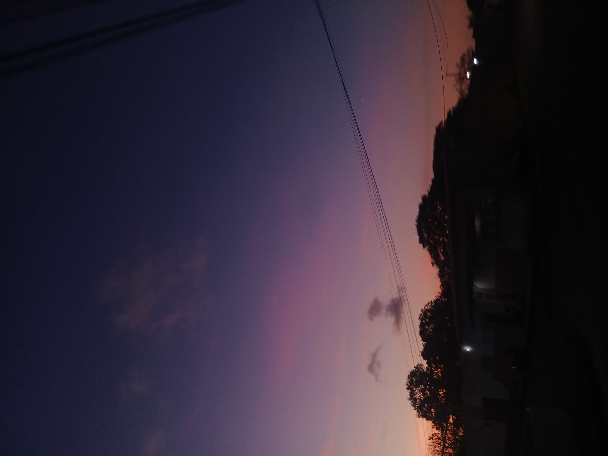 The image depicts a dusky scene in what is possibly Juan Griego, Venezuela. The sky exhibits a gradient of colors, transitioning from deep purplish-blue on the left to a blend of orange and pink hues on the right. Power lines are visible against the sky. Along the lower right edge of the image, there are dark silhouettes of trees and buildings. Lights can be seen within the buildings. The right side of the image is obscured by darkness, possibly indicating the edge of a street or road.