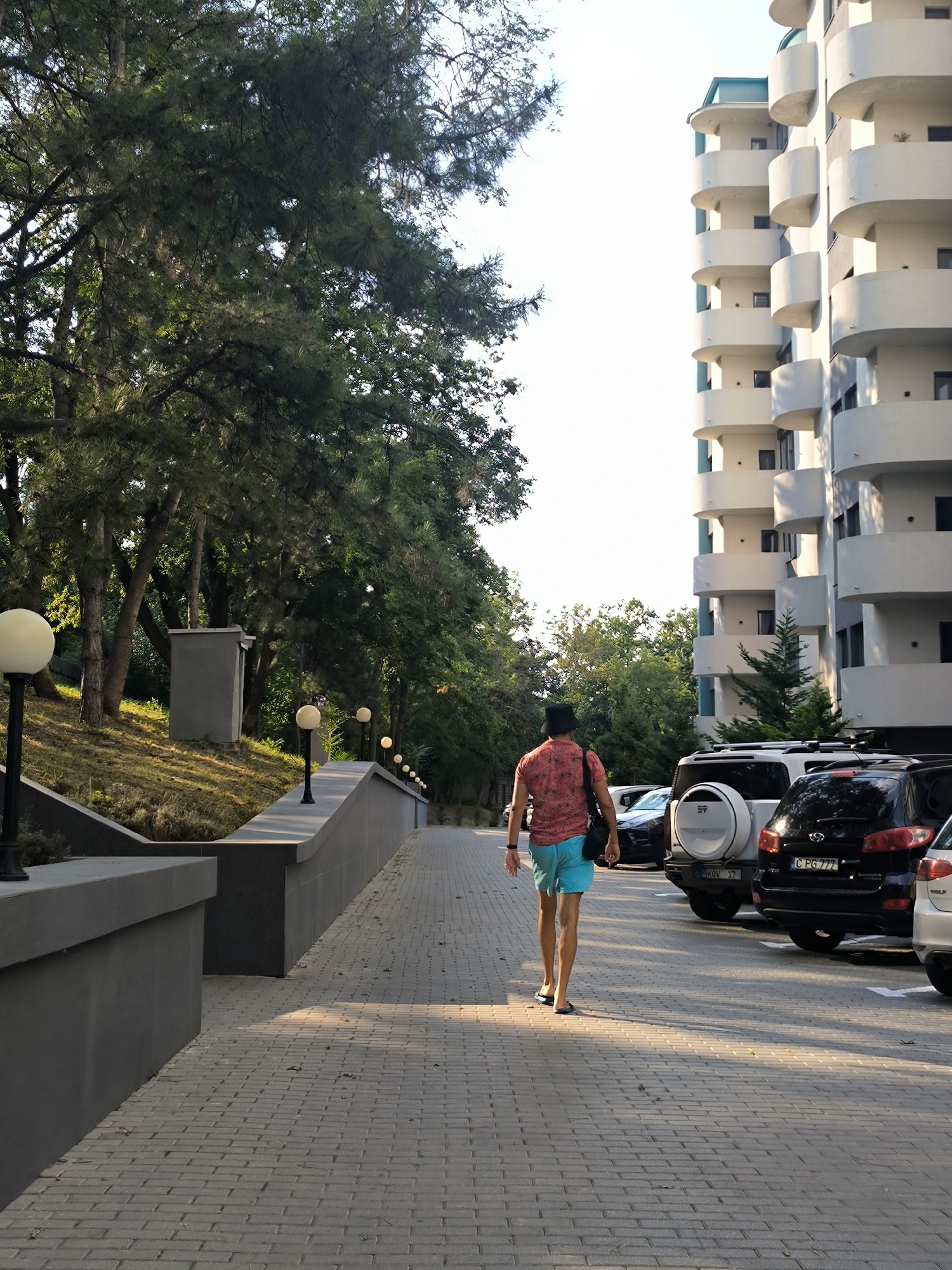 A man wearing a patterned short-sleeve shirt and blue shorts, with a black bag slung over his shoulder and a black hat on his head, walks away from the camera on a brick-paved walkway. He is on the right side of the frame, moving towards a line of parked cars. To the left of the walkway is a grey raised wall with lights mounted on it, and beyond that, a grassy embankment with trees. In the background, a modern, multi-story white building with rounded balconies is visible. The scene appears to be in a residential area, possibly near a park or a beach, given the greenery and the building's design. The lighting suggests it is daytime, with shadows cast by the trees and walkway features. This image was taken in the city of Ustia, Moldova.