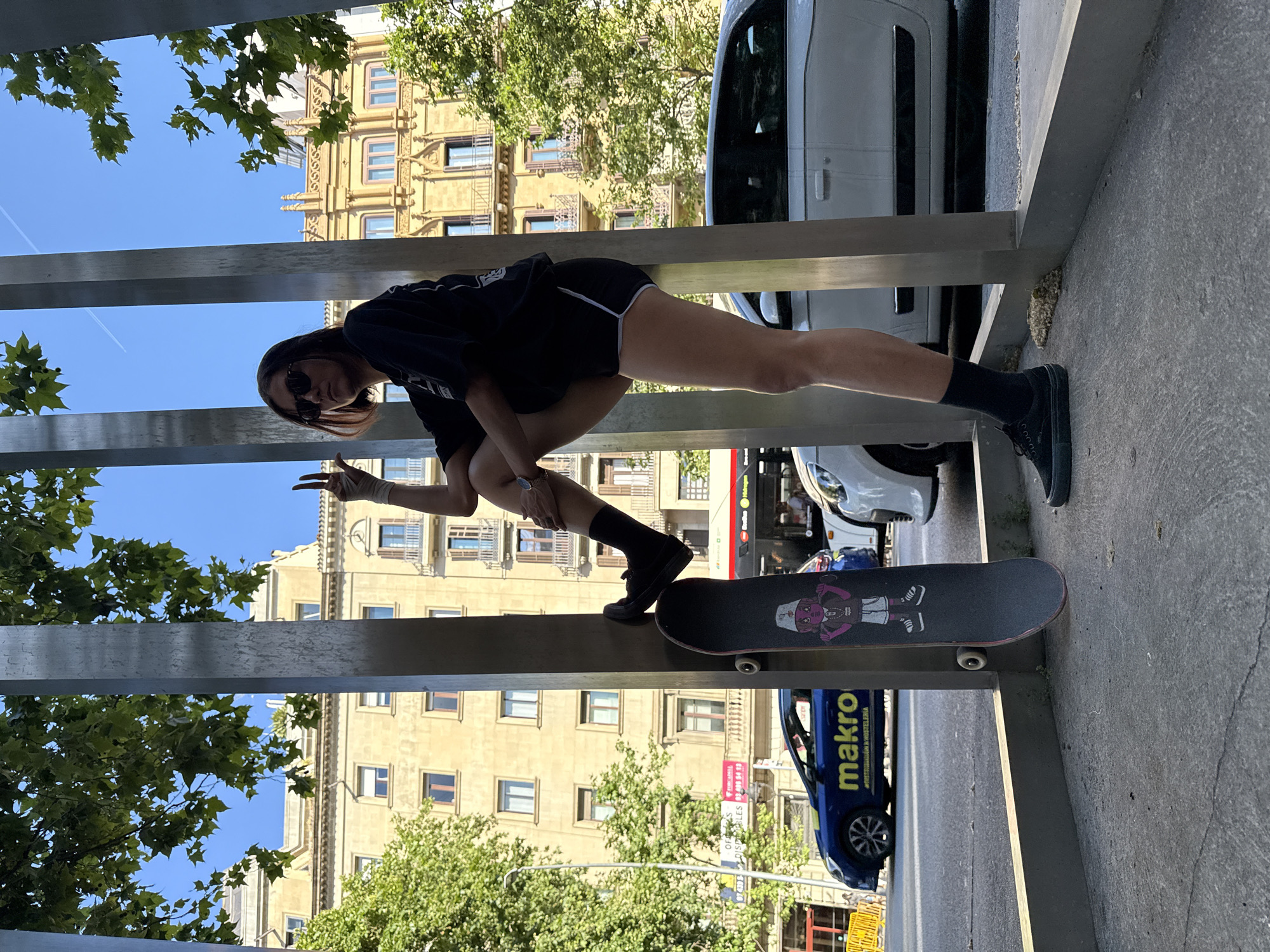A young adult female stands on a grey skateboard atop a grey architectural beam structure. The female wears a dark shirt, dark shorts with a white stripe, dark socks, and dark sneakers. Her right hand is wrapped, and she is displaying a peace sign. Behind the structure, a light brown building with windows is visible. Vehicles, including a white van and a blue car with the word "makro" on it, are partially seen in the background. Foliage from trees is present on both sides of the structure. The scene appears to be in an urban environment, likely Barcelona, Spain, given the presence of "makro" advertising and the architectural style. The image is rotated counterclockwise.
