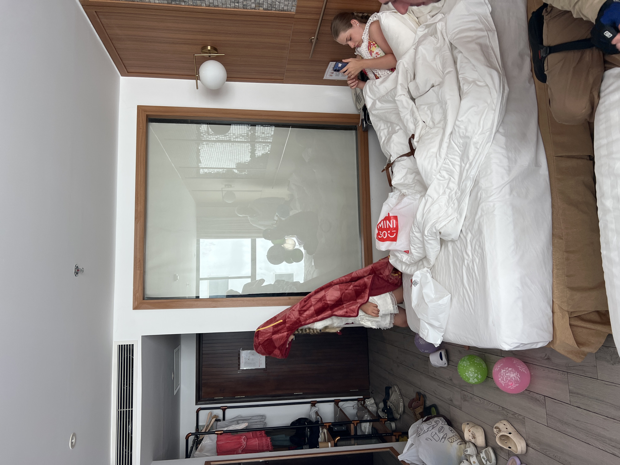The image depicts a hotel room with two young girls. One girl, wearing a white dress and a red hooded cape, stands near a wooden door and a clothing rack. The other girl, lying in a bed covered with white linens, uses a smartphone. A large window with a reflection of other people and objects dominates the background. Several items are scattered on the wooden floor, including shoes, bags, and balloons. The room features a large bed, a wooden wardrobe, and modern lighting fixtures. The overall impression is one of a casual and possibly playful atmosphere within a hotel setting.