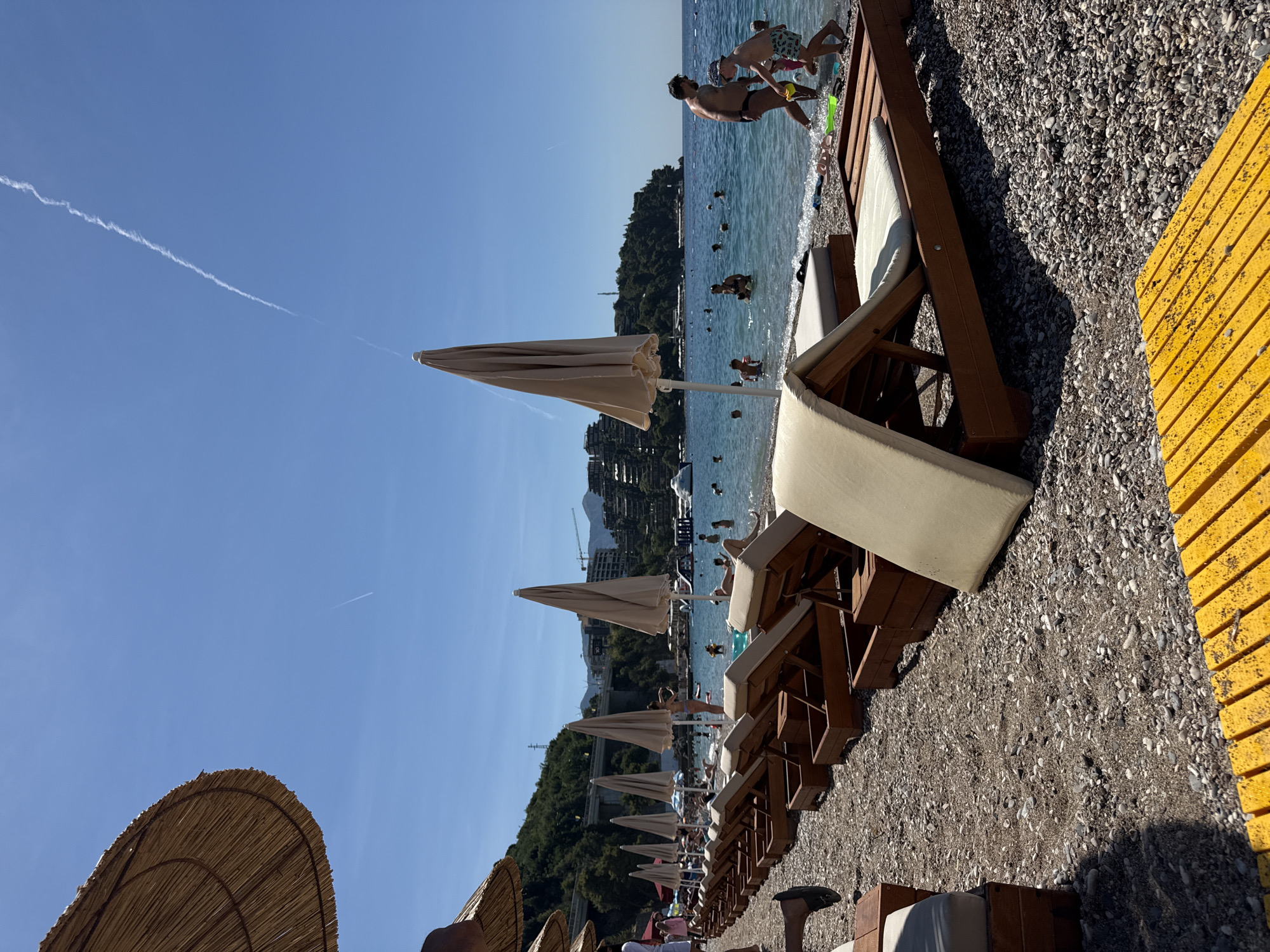 The image depicts a busy beach scene on a clear, sunny day in Boreti, Montenegro. In the foreground, rows of empty, wooden lounge chairs with beige cushions are set up on a pebble beach. Some of these chairs are partially covered by large, straw beach umbrellas, and one straw umbrella is prominently visible in the upper left corner. A bright yellow, slatted walkway runs along the bottom of the frame.
In the middle ground, the clear, turquoise water of the Adriatic Sea is populated with numerous people swimming and wading. Some individuals are visible on the beach near the water's edge. A father and son are walking out of the water, the son carrying a green bucket. Further out, a family with children plays in the shallow water. Various inflatable toys can be seen floating.
The background features a hilly, tree-covered coastline with buildings and structures built into the hillsides. A bridge or elevated roadway is visible crossing over the land. The sky is a vibrant blue with a thin, white contrail from an aircraft stretching across it. The overall impression is one of a well-established tourist beach location, with ample amenities for visitors.