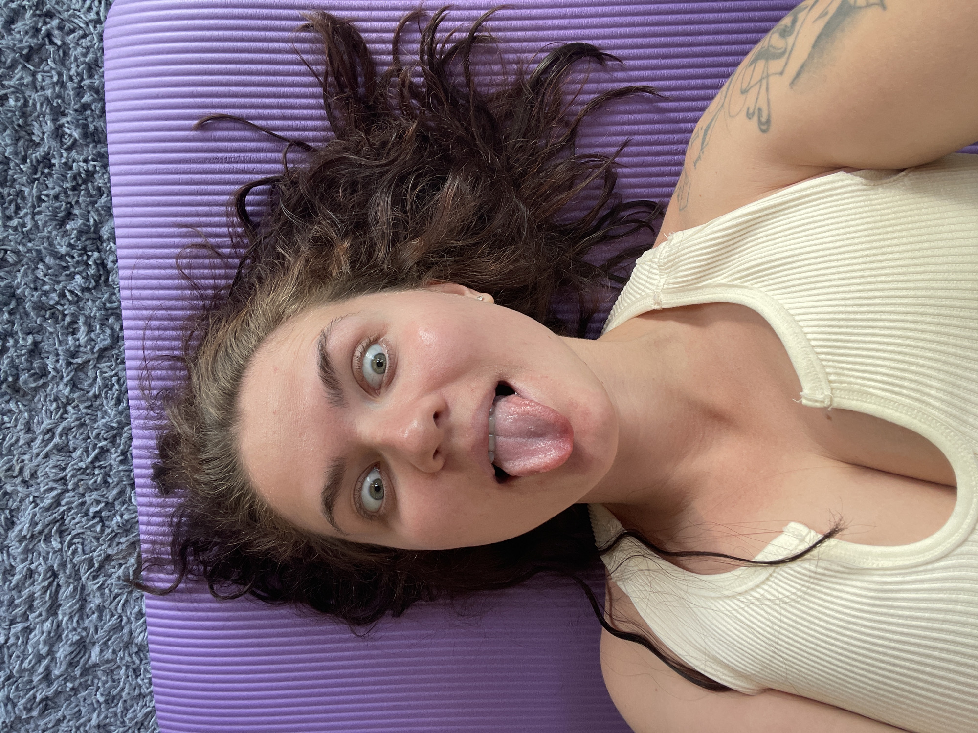 A woman with fair skin, curly brown hair, and light green eyes is lying on a purple yoga mat on a gray carpet. Her hair is spread out around her head. She is wearing a white tank top. Her mouth is open, and her tongue is sticking out. A tattoo is visible on her upper arm. The location is not identifiable as Мосметаллоконструкция Онт, Russia from this image.