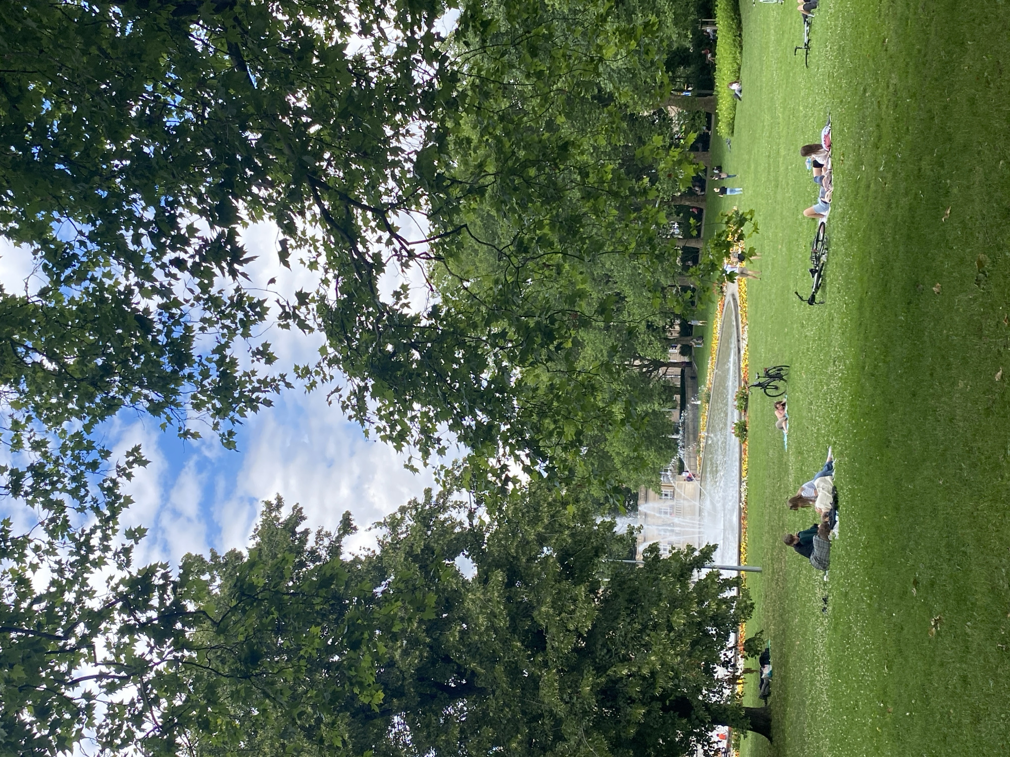 This image captures a serene park scene in Poznan, Poland, likely within the area of the Freedom Square (Plac Wolności) or a similar large green space. In the background, a large fountain with multiple streams of water is active, with a classical building visible behind it. The foreground is dominated by a lush green lawn dotted with white clover flowers. Several people are scattered across the lawn, enjoying the outdoors.
In the middle ground, two individuals are seated on a blanket, appearing to be in conversation. To their right, another person lies on a blanket, resting. Further to the right, a bicycle is parked near a person reclining on a blanket. In the distance, several more individuals are visible, some walking and others appearing to relax on the grass. The park is framed by the dense foliage of mature trees, their leaves creating a dappled effect as they filter the sunlight and frame the sky, which is a mix of blue with scattered white clouds. The overall atmosphere is peaceful and recreational, suggesting a pleasant day for outdoor leisure in Poznan.