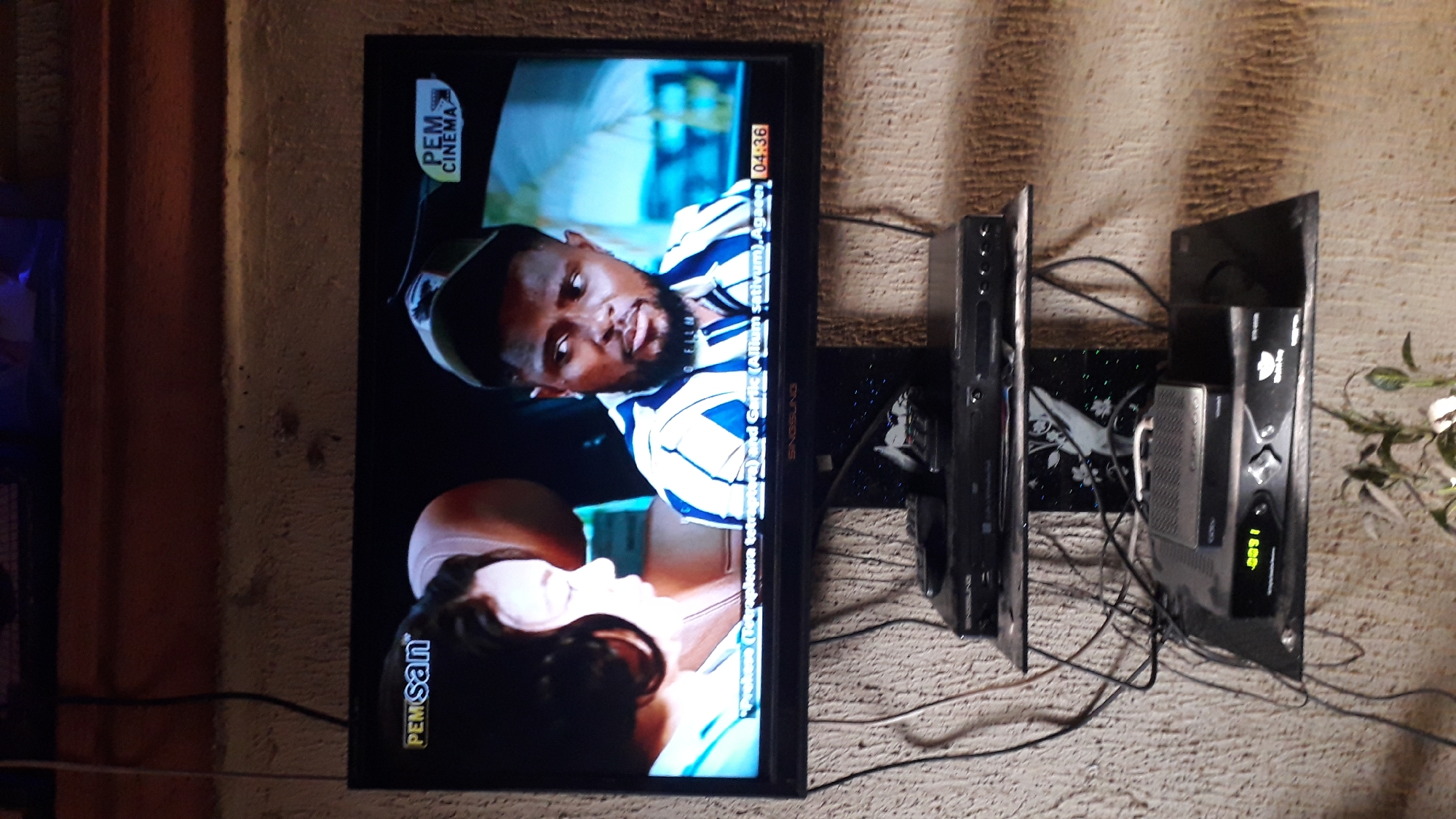 The image depicts a television screen displaying a scene from a movie or TV show. The screen shows two individuals, a woman on the left and a man on the right, who appear to be engaged in a conversation. The man is wearing a blue and white striped shirt and a cap. The television is mounted on a wall in what appears to be a residential setting. Below the television, there are two shelves holding electronic devices, including what look like DVD players and a set-top box. Several cables are visible, connecting these devices. The wall behind the television and shelves has a textured finish. The setting is likely a home in Nkpor, Nigeria, judging by the context of the request. The overall scene is domestic and everyday.