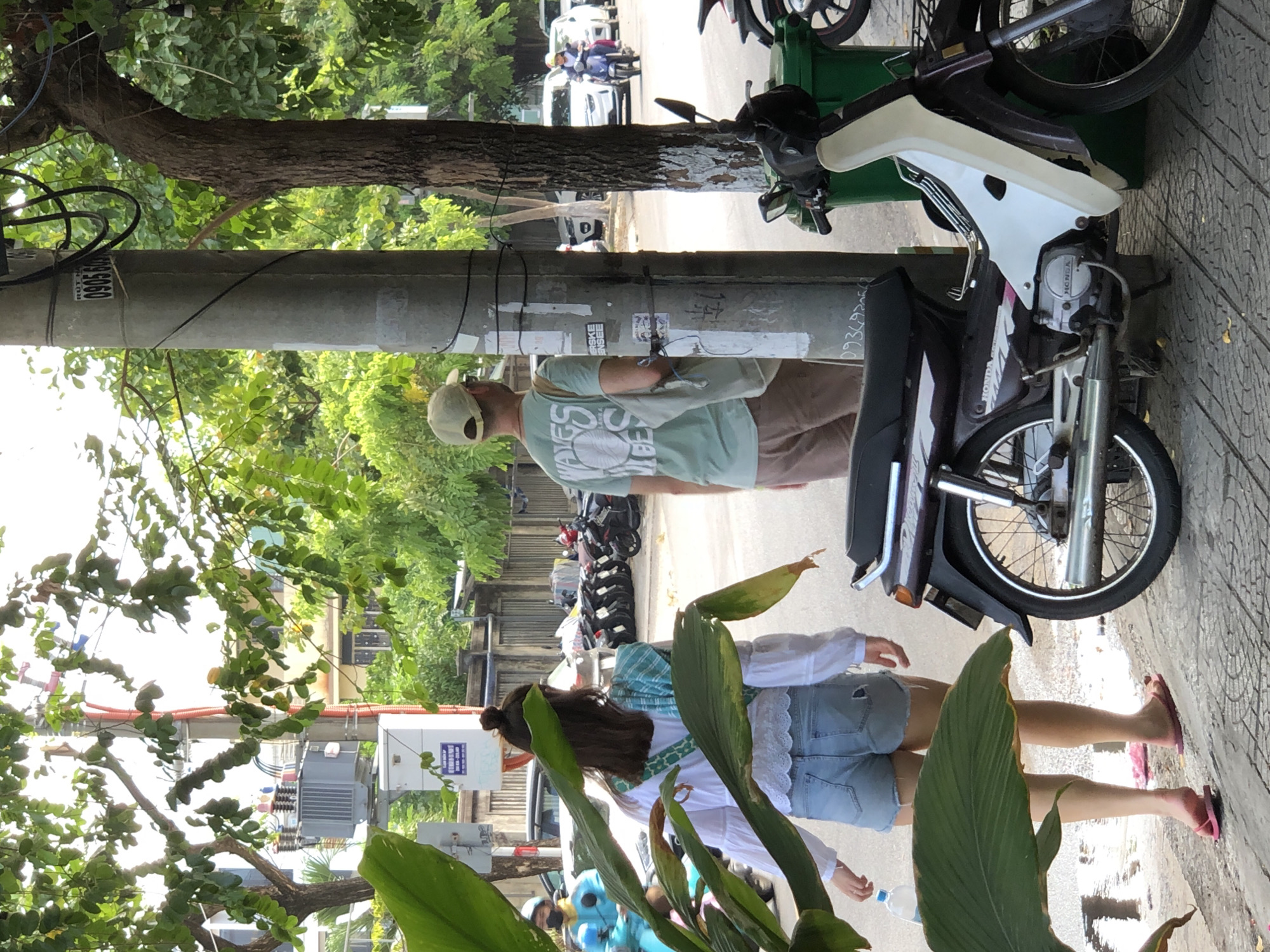 The image captures a street scene in Đà Nẵng, Vietnam. In the foreground, a woman with dark hair tied in a bun, wearing a white long-sleeved shirt and denim shorts, walks away from the camera. She carries a clear plastic water bottle in her left hand and a light blue and green checkered fabric is draped over her right shoulder. Her feet are bare, wearing pink flip-flops. To her right, a man in a light green t-shirt with a graphic design and a light beige baseball cap stands facing away from the camera, holding a beige tote bag over his left shoulder. He wears beige shorts. A tall, gray utility pole stands between the woman and the man, adorned with stickers and cables. Partially visible to the right of the pole is the front of a dark purple and white motorcycle. In the background, a line of parked motorcycles is visible, along with white cars on the street. A person in a bright blue uniform and helmet, likely riding a motorcycle, is seen on the far left side of the image. Lush green trees with dense foliage overhang the scene, casting shadows and contributing to the vibrant atmosphere of a tropical urban environment.