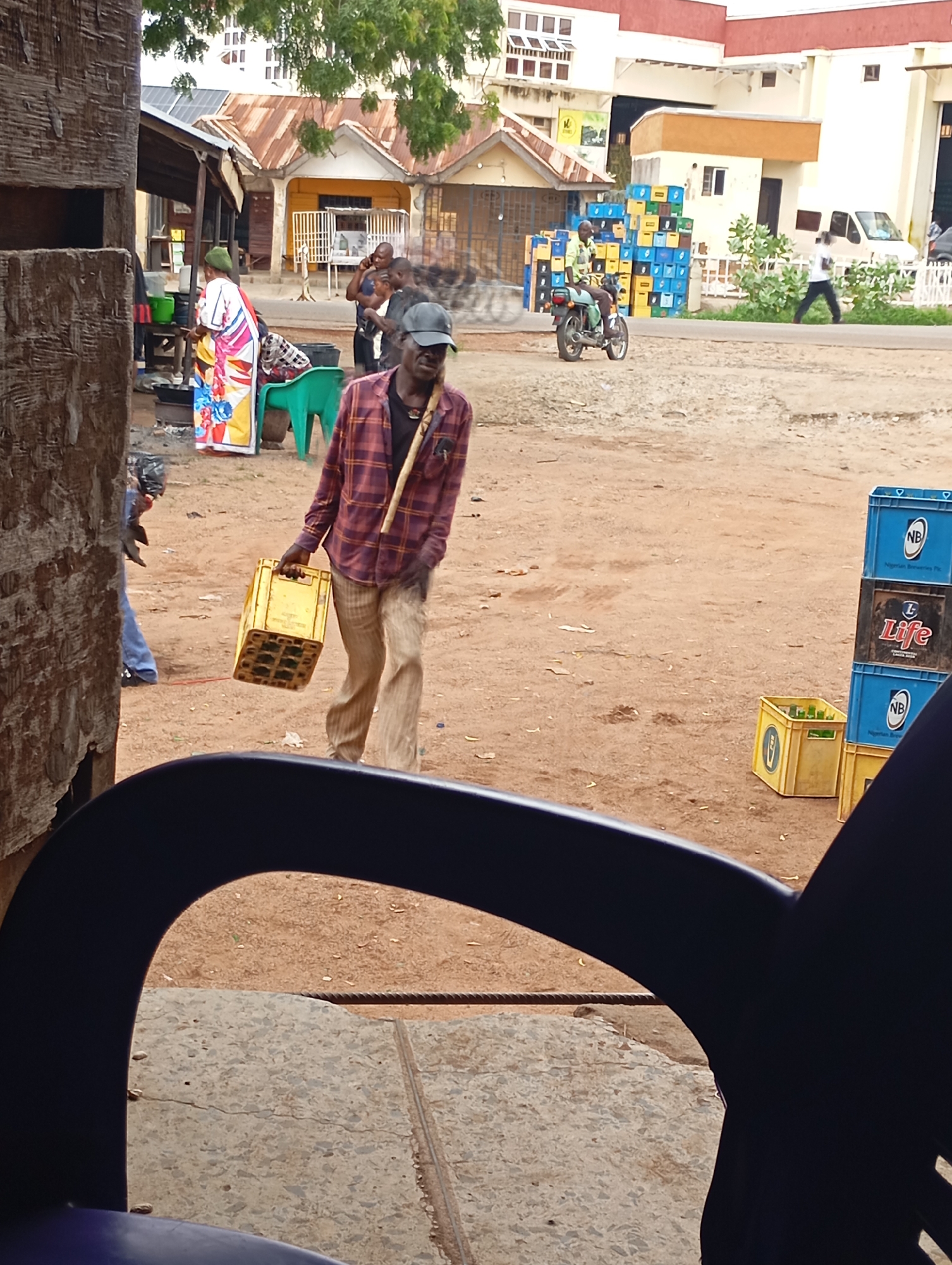 The image shows a street scene in Shendam, Nigeria. In the foreground, a man wearing a plaid shirt and light-colored pants is walking away from the camera, carrying a yellow crate filled with bottles. He has a dark baseball cap on and a stick over his shoulder. Behind him, a woman in a colorful dress and headscarf is standing near a stall. Further in the background, there are stacks of blue and yellow crates, some with "NB" and "Life" labels, suggesting they contain beverages. A motorcycle is parked nearby, and other people are visible in the distance. The ground is dusty and unpaved. Buildings with corrugated iron roofs are present, indicating a commercial or market area. A white van is parked on the right side of the image. The overall scene depicts everyday life and commerce in an outdoor setting.