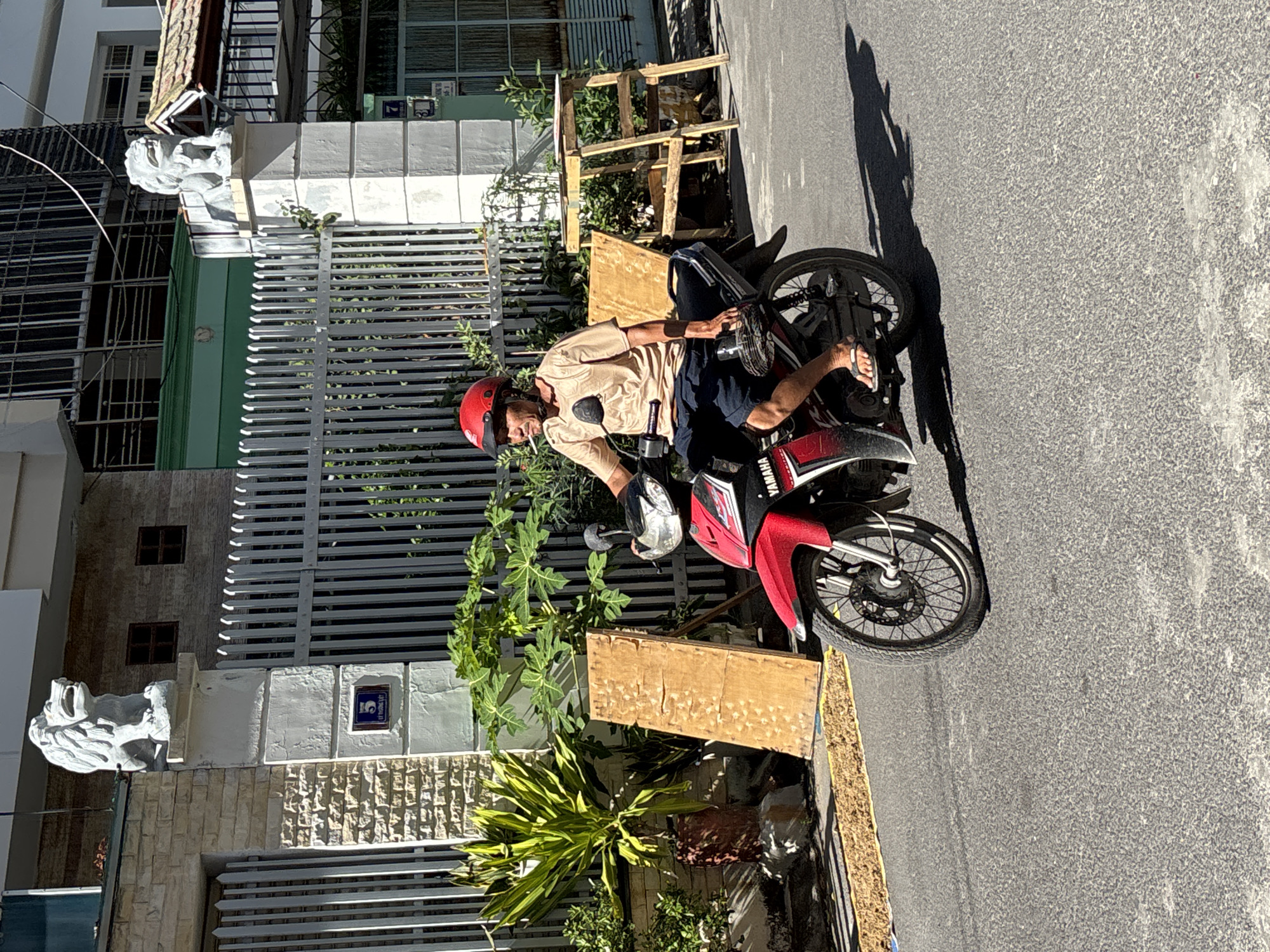 A man in a tan shirt, dark shorts, and a red helmet is riding a red and black Yamaha motorcycle on a street in Nha Trang, Vietnam. He is smoking a cigarette. The motorcycle has a prominent "YAMAHA" logo on its side. Behind him, a tall metal fence separates the street from a property with a brick wall, a large leafy plant, and what appears to be a wooden plank leaning against a fence post. Further in the background, there's a building with windows and a green awning. Two lion statues are visible on top of pillars in front of the property, and a sign with the number "5" is affixed to one of the pillars. The street surface is asphalt. The scene is illuminated by bright sunlight, casting shadows.