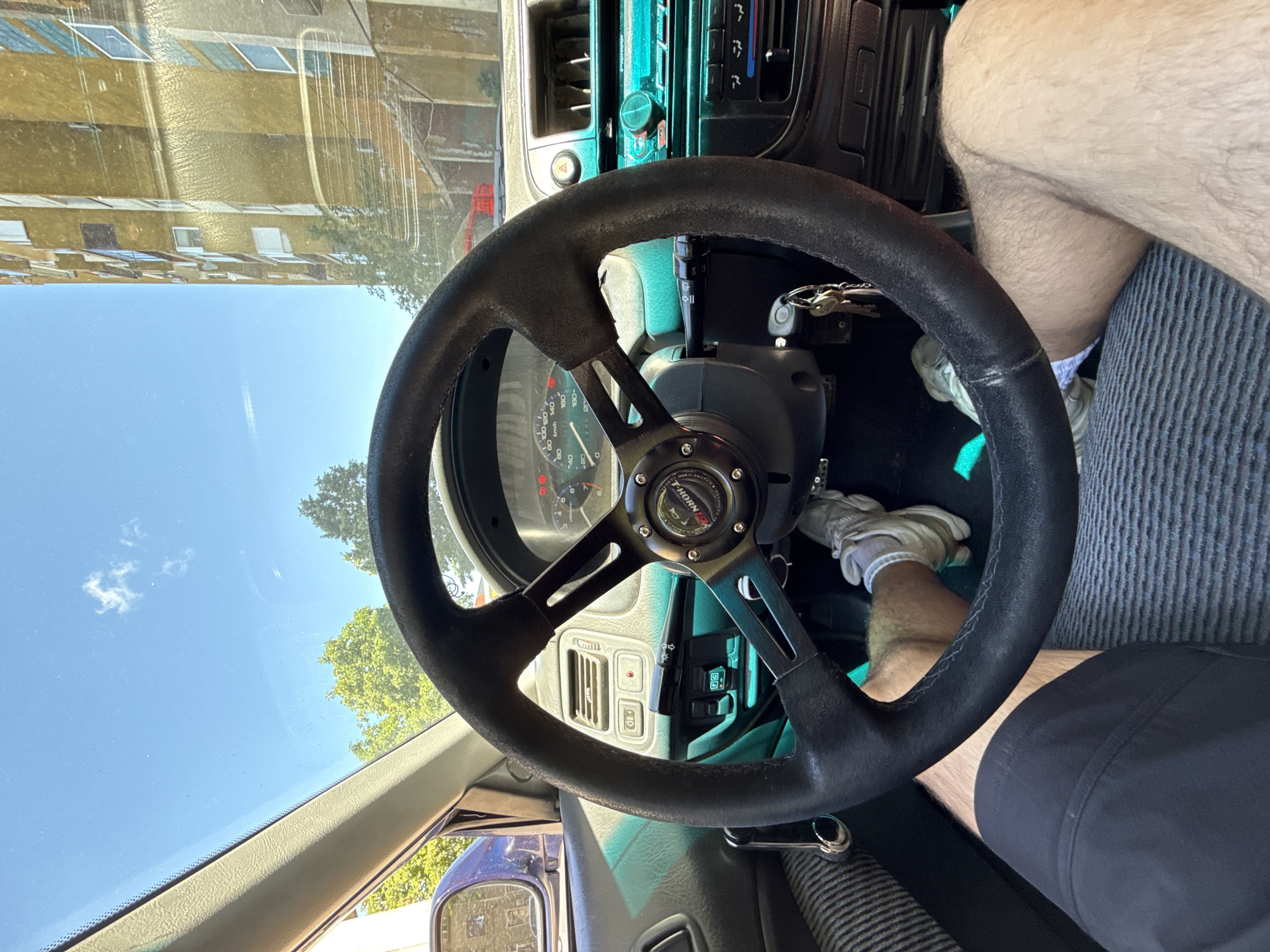 The image shows the interior of a car, looking forward from the driver's seat. The steering wheel is prominent in the foreground, with a person's legs and feet visible. The dashboard features gauges, a radio, and air conditioning controls. Outside the car, through the windshield, a bright blue sky with a few clouds is visible, along with trees and a tall building. The scene appears to be a sunny day in an urban environment. No specific location in Shumen, Bulgaria can be identified from the image.
