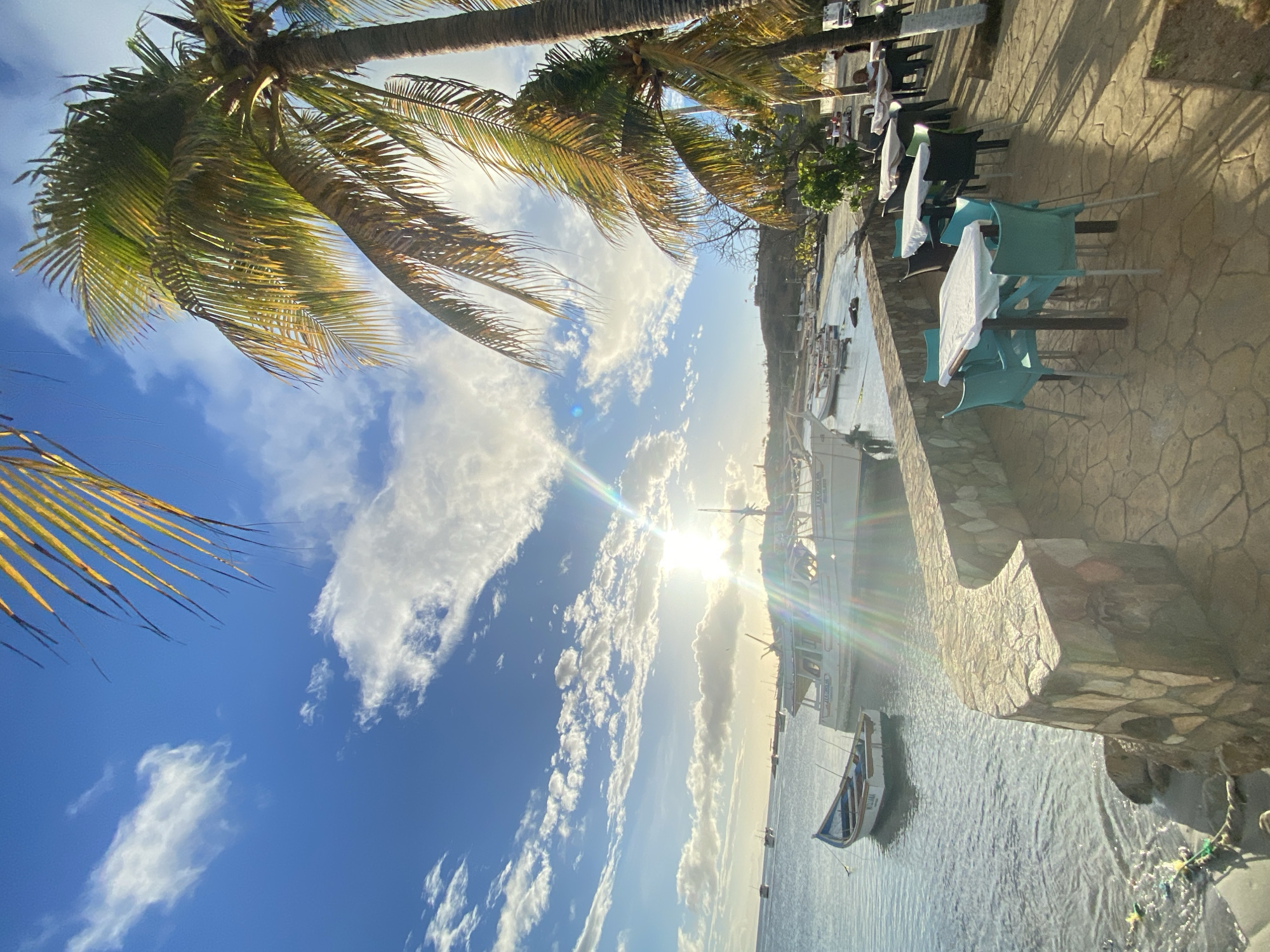 The image captures a waterfront scene in Juan Griego, Venezuela, bathed in sunlight with a bright glare near the horizon. Palm trees frame the upper and left sides of the image, their fronds reaching into a sky filled with scattered clouds. To the right, a stone wall separates a dining area with tables and light-blue chairs, some covered with white cloths, from the water. Several boats are visible in the water, including a small white-and-blue rowboat closer to the foreground and larger boats further out.