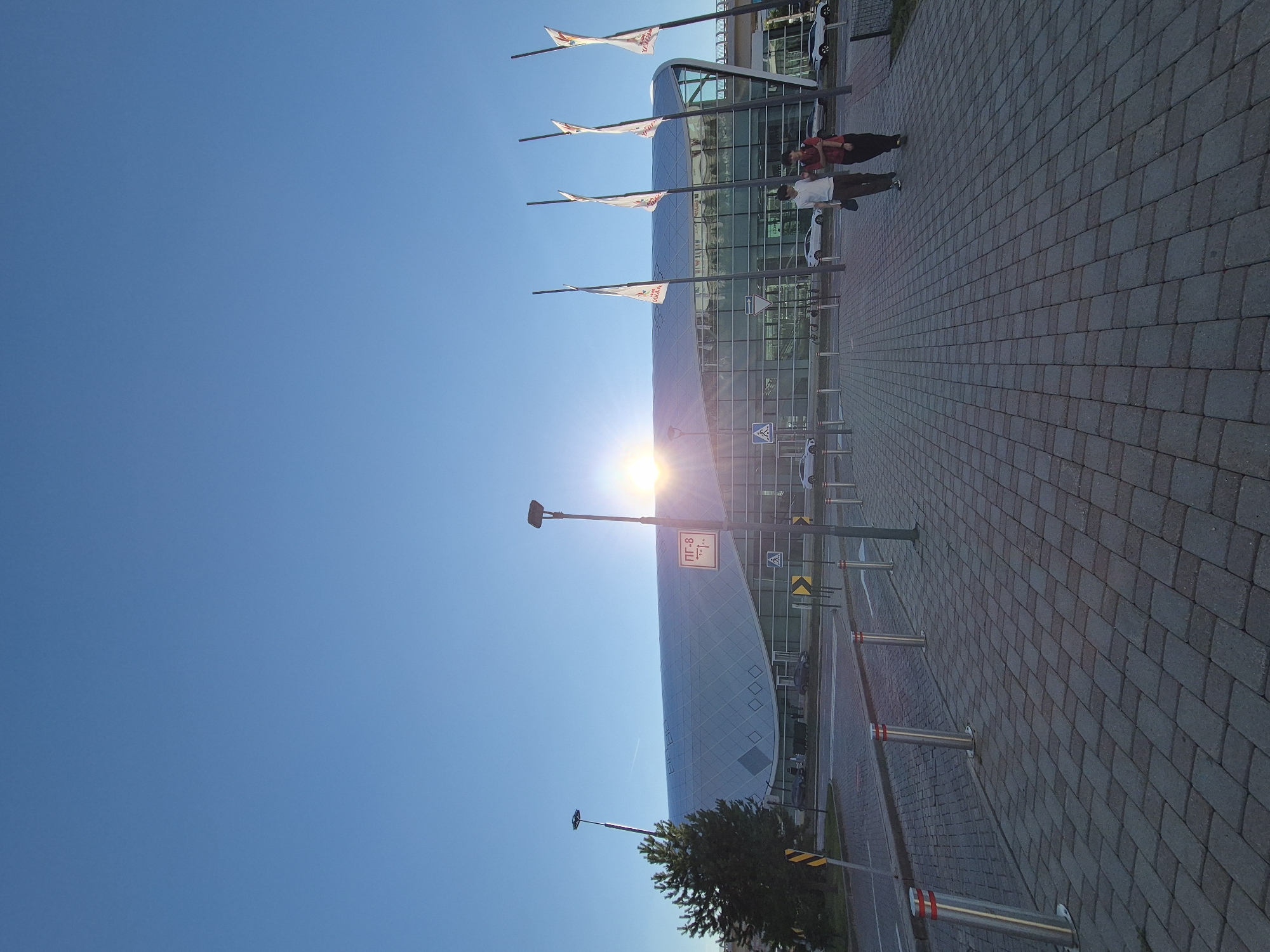 The image depicts the modern architectural facade of Nursultan Nazarbayev International Airport in Astana, Kazakhstan, under a clear blue sky. The sun is low on the horizon, casting a lens flare that is prominently visible. The airport building features a curved, glass-fronted design with a patterned roof. Several white flags with red logos are planted in front of the building. A paved walkway made of interlocking grey bricks leads towards the airport entrance. Along the walkway are bollards with red reflective bands. Two individuals, appearing to be young men, are walking on the walkway away from the camera. One is wearing a white t-shirt and dark trousers, and the other is wearing a red t-shirt and dark trousers. Several cars are visible parked or driving in the background. A traffic light and directional signs are also present. A lone evergreen tree stands to the left of the image.