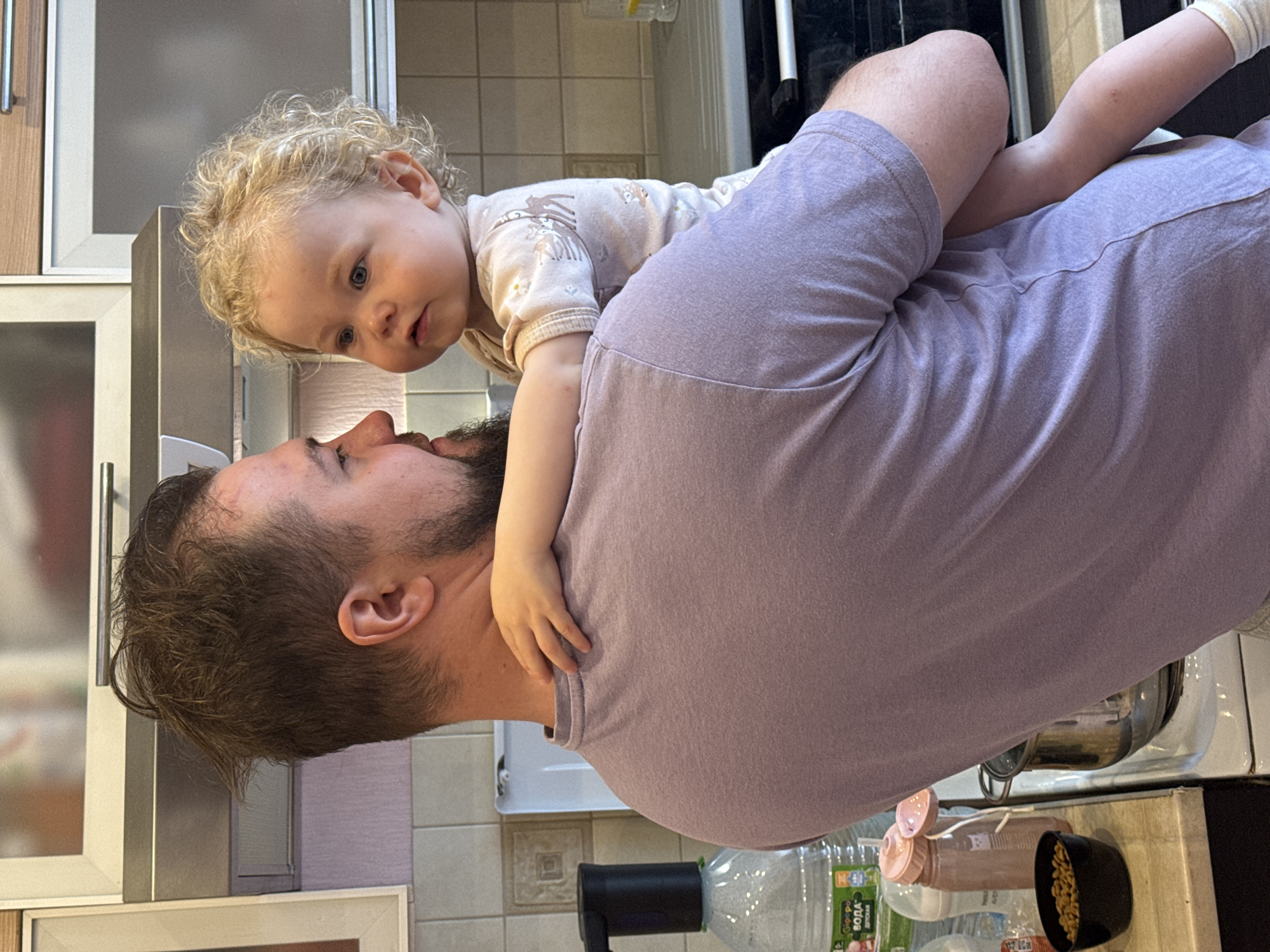 This image captures an affectionate moment between a man and a baby, likely a father and daughter, in a home kitchen setting in Vladimir, Russia. The image is oriented vertically, with the man positioned in the foreground, his back largely to the viewer, but his head turned to face the baby.
The man has dark brown hair and a beard and is wearing a light purple t-shirt. The baby, with curly blonde hair and bright blue eyes, is situated on his shoulders or upper back, leaning forward to look directly into his face. Her small hands are gently resting around his neck or shoulder. She is dressed in a light-colored, long-sleeved onesie patterned with subtle animal and floral designs. The baby's expression conveys curiosity and engagement, while the man's partially visible profile suggests a tender and playful interaction.
The background shows elements of a domestic kitchen. There are light-colored tiled walls, possibly serving as a backsplash, and light-toned kitchen cabinets, some featuring frosted glass inserts. On a dark countertop visible in the lower right, several items are present: a large clear plastic bottle with a black pump dispenser, a smaller pink bottle (resembling a baby bottle or sippy cup), and a black bowl filled with small, light-colored contents, possibly seeds or nuts. Visible text on the large water bottle's label reads "БЮВЕТ ВОДА ДЕТСКАЯ," which translates to "BÜVET BABY WATER." The scene depicts a warm, indoor interaction, likely occurring during daylight hours.
