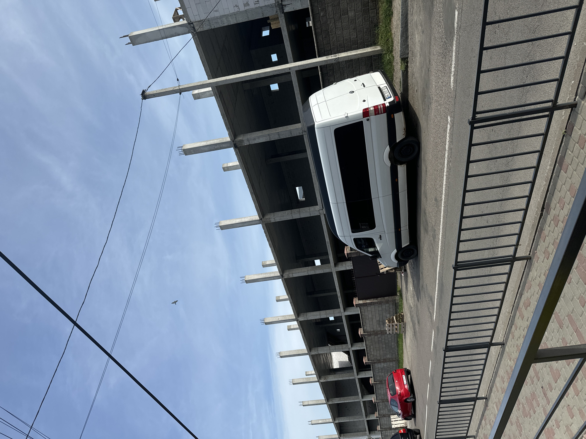 The image depicts an outdoor scene featuring a multi-story building under construction, a white van parked on the street next to it, and a red car further down the road. The building's concrete framework is visible, with exposed rebar and unfinished floors. Overhead power lines crisscross the sky, and a bird is captured in flight against the blue sky. The street is paved and marked with white lines, and a black metal fence runs along the sidewalk. The setting appears to be a daytime scene with clear weather. The scene is likely in an urban or semi-urban environment, given the construction and road infrastructure. No people are visible in the image. The license plate of the white van is partially visible and appears to be Ukrainian. The overall impression is one of ongoing development or urban construction.