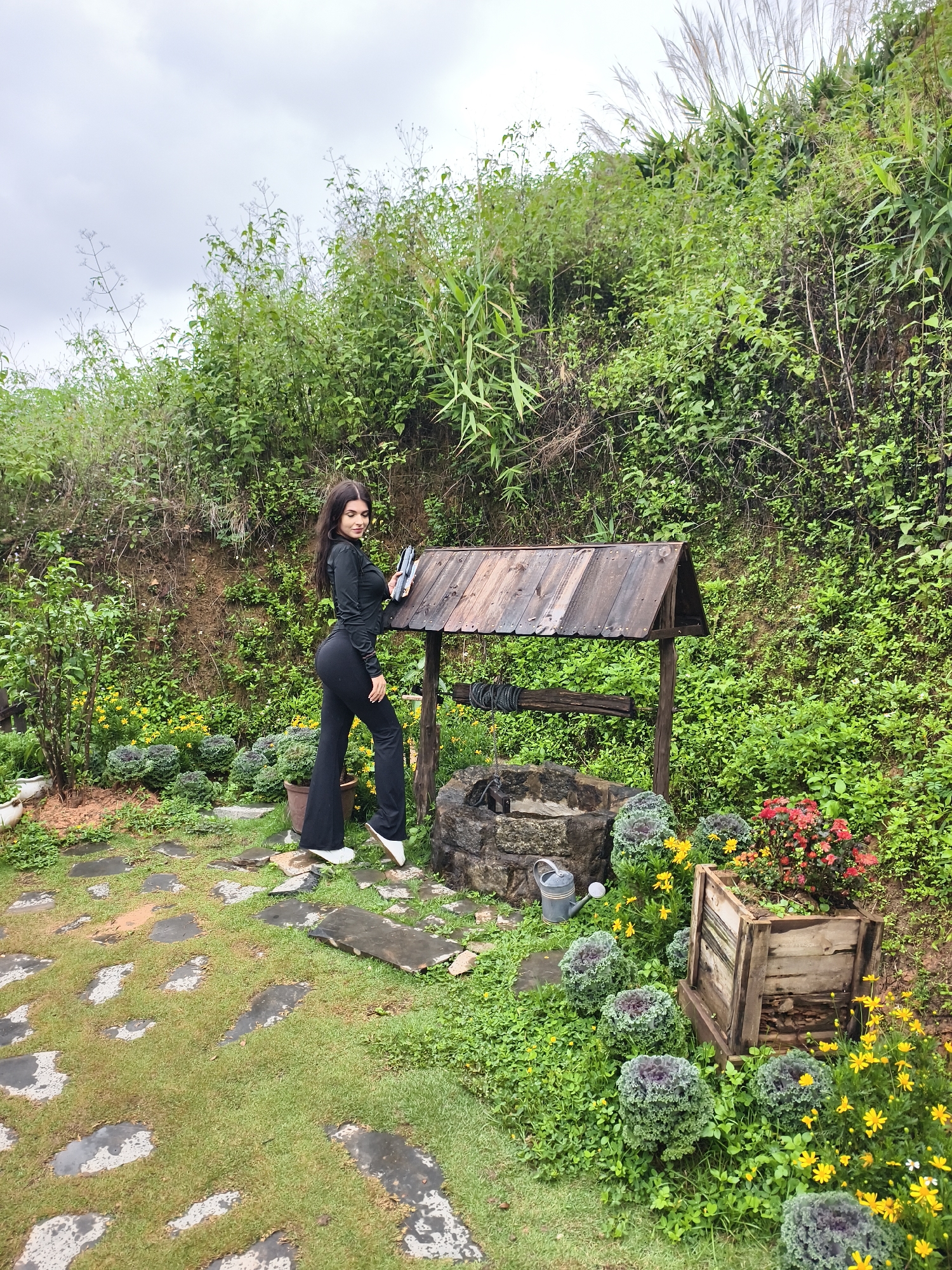 A young woman in a black outfit and white shoes stands near a wishing well in Thôn 1, Vietnam. The well is made of stone and wood with a small roof. Lush green vegetation surrounds the well. The ground is covered in grass and stone pathways. There are flowers in wooden crates, and a watering can sits nearby. The sky is overcast.