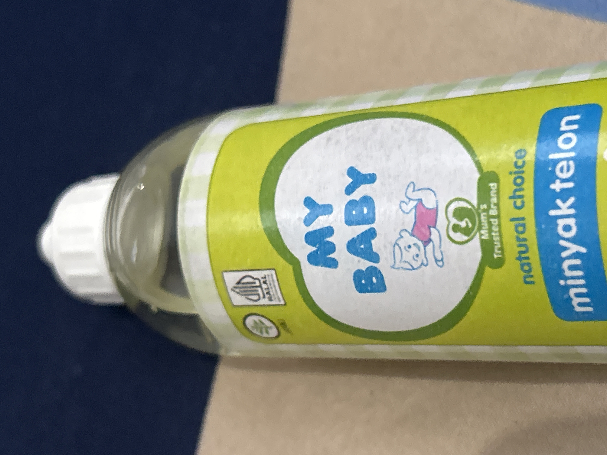 A clear plastic bottle of "MY BABY minyak telon" (telon oil) is depicted horizontally, with its white screw cap pointing to the left. The bottle contains a light yellow liquid, typical of telon oil. Its label is predominantly light green with a darker green oval border, featuring the brand name "MY BABY" in prominent blue letters within the oval. A cartoon image of a baby, wearing a pink top and blue shorts, is lying beside the text. Below the baby image, a small green circle contains the number "3" and the text "Mum's Trusted Brand." To the right of the oval, the words "natural choice" are visible in green, followed by "minyak telon" in a blue rectangular box. On the far left of the label, a white "HALAL" certification logo, presented in both Arabic script and Latin letters, is visible. Below the HALAL logo, there's a small green circular symbol, possibly indicating a natural ingredient or eco-friendly aspect. The background consists of a dark blue surface on the left and a light brown or tan textured surface on the right, suggesting the bottle is resting on a surface that contrasts in color and material, possibly a bed or table. There is no specific activity taking place, as it's a still image of a product. The image provides no cues for the time of day, weather, or emotions, but the product type and "HALAL" certification align with the given location context of Lhokseumawe, Indonesia, where such products are commonly used.