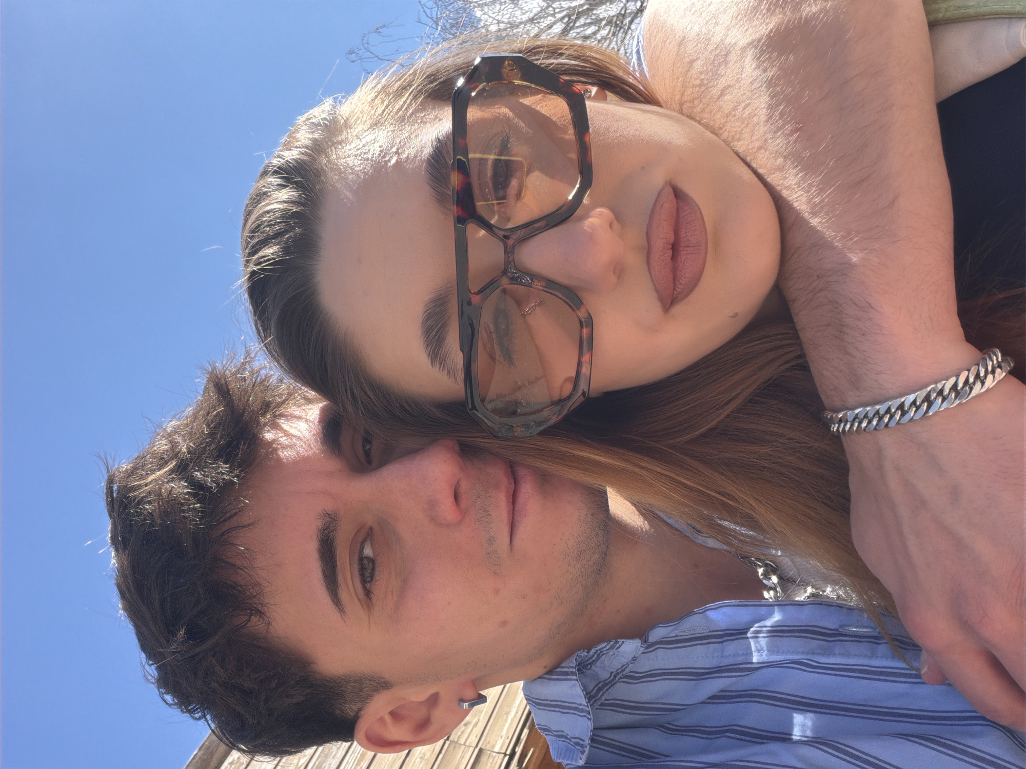 A selfie image depicts a man and a woman outdoors. The man, with dark hair and a blue striped shirt, is at the bottom left. The woman, with long brown hair and sunglasses, is above him. Her arm, adorned with a silver bracelet, is around him. The background shows a blue sky. The location is possibly Sofia, Bulgaria, but there are no specific landmarks to confirm this.