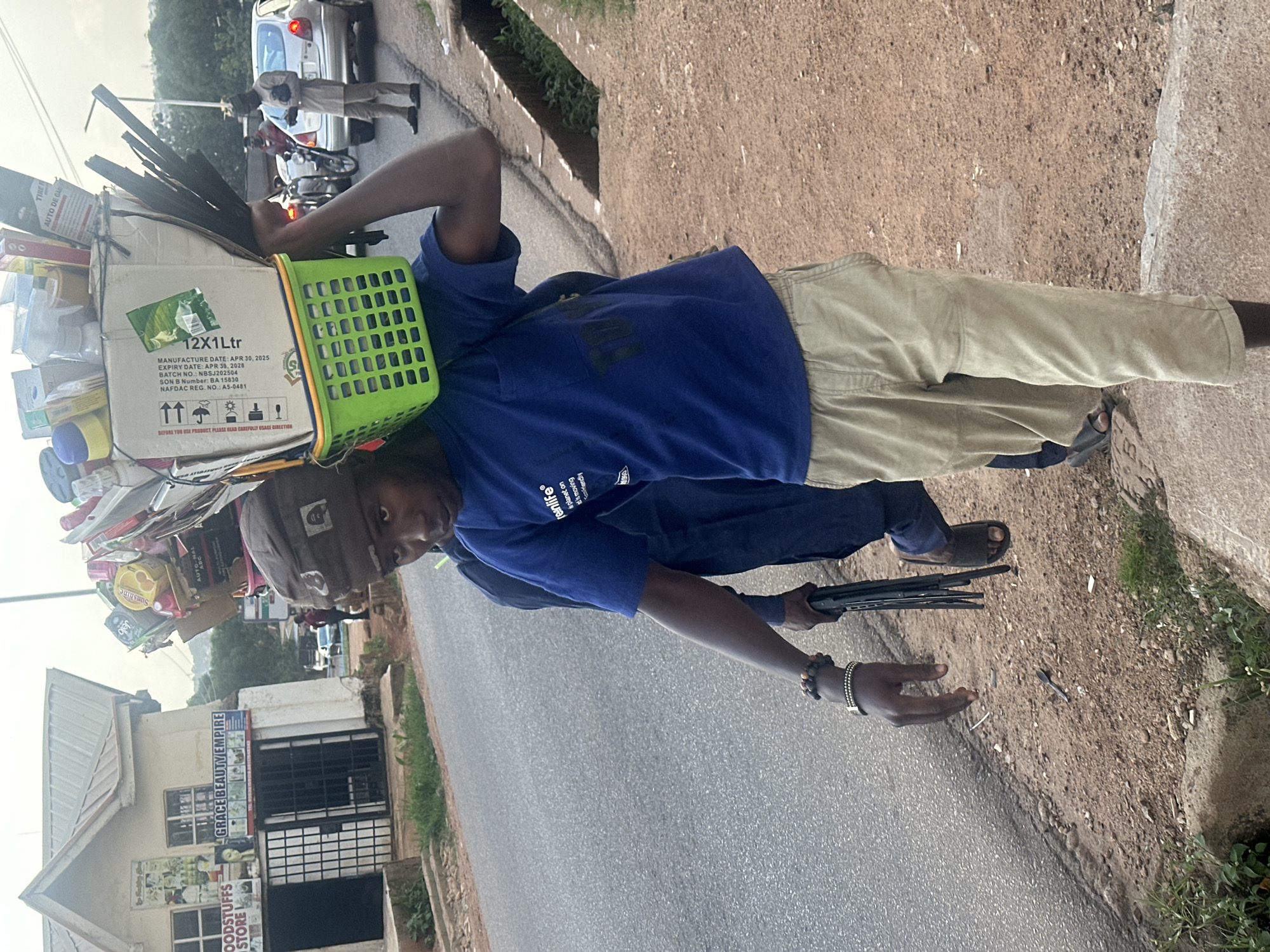 A young male, appearing to be in his late teens or early twenties, walks along a dusty sidewalk on the outskirts of Gura Top, Nigeria, carrying a large basket filled with various items, balanced on his head. He is wearing a blue t-shirt with white lettering that reads "re.inlife" and tan trousers. His left hand is adorned with several beaded bracelets and a watch, and he is holding a black object, possibly a foldable umbrella or cane, in his right hand. Another individual, partially visible behind him and wearing a dark blue garment, walks in the same direction.
The background reveals a street with a paved road, a drainage ditch, and a building on the left side of the image. The building has signage that reads "GOODSTUFFS STORE" and "GRACE BEAUTY EMPIRE." The environment suggests a typical urban or peri-urban setting in Nigeria, characterized by informal businesses and busy roadways. Cars and a motorcycle are visible on the road in the middle ground, indicating traffic. The overall scene depicts everyday life and commerce in the region.