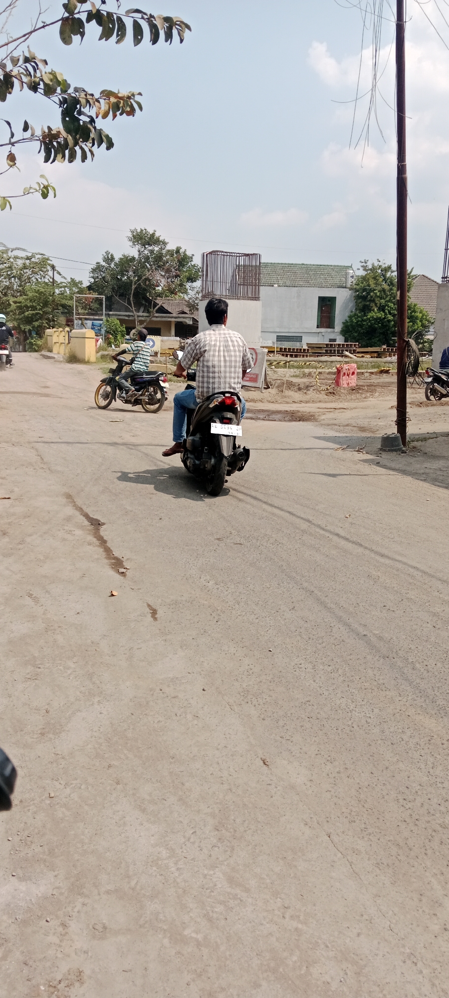 The image depicts a scene on a paved road in Purwomartani, Indonesia, under a bright, slightly hazy sky. In the foreground, a person wearing a checkered shirt and blue jeans is riding a black motorcycle, facing away from the camera. The motorcycle has a license plate with the inscription "SB 3434 ZH". To the left of this motorcycle, another person on a darker motorcycle with a checkered shirt and dark pants is also riding away from the camera. In the background, there is a white building with a green tiled roof, and construction materials are visible, suggesting ongoing development. Trees and vegetation are present on either side of the road, and a utility pole stands on the right side of the frame. The road surface is concrete with visible tire marks and some debris.