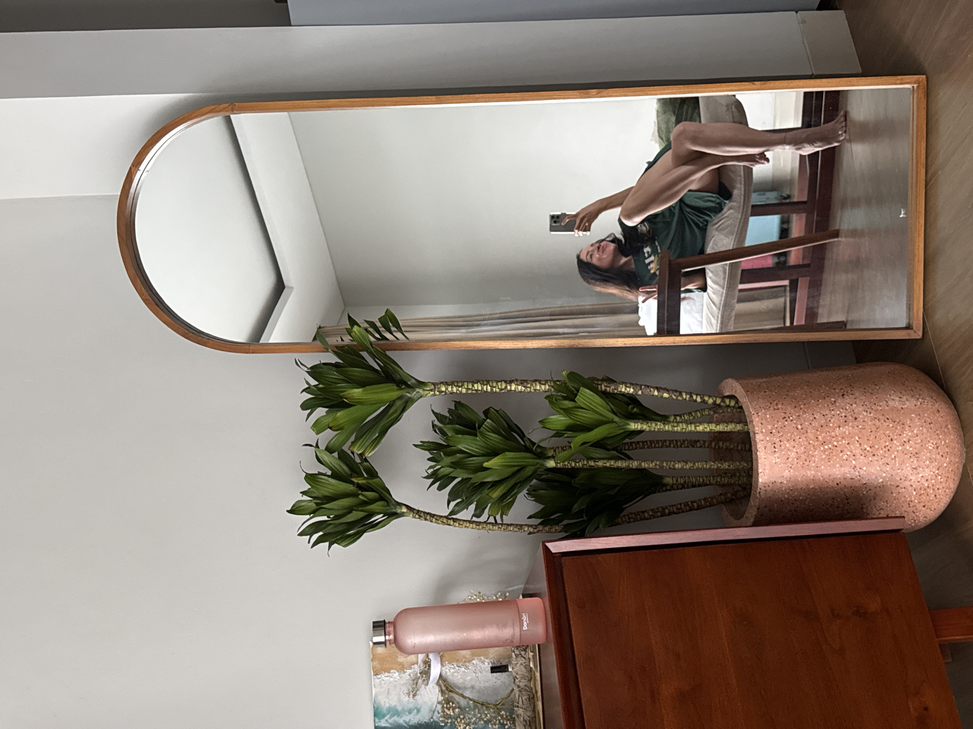 The media file shows a full-length mirror with a wooden frame, reflecting a woman sitting on a chair. The woman is taking a selfie with her phone. She is wearing a green top and dark shorts. The room appears to be a bedroom or living area, with a large plant in a speckled pink pot to the left of the mirror. A wooden dresser or table is partially visible in the foreground. The scene suggests a casual, indoor setting. The lighting indicates it is daytime, possibly morning or afternoon. The overall mood is relaxed and personal, as evidenced by the selfie. The location context is North Kuta, Indonesia. There is no visible weather information. There is text on the pink water bottle, but it is not clearly legible.