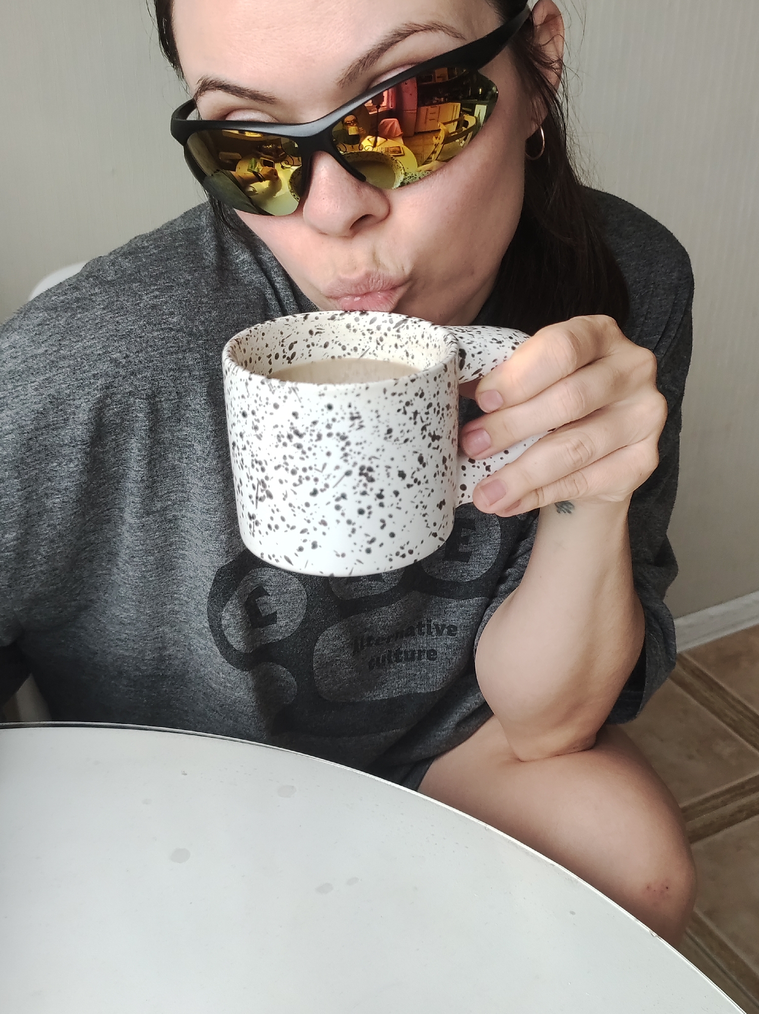 A medium shot depicts a person with dark hair and sunglasses puckering their lips while holding a white mug with black speckles. The mug appears to contain a light-colored liquid, possibly coffee or tea. The person is wearing a gray shirt with a design. The reflection in the sunglasses suggests the person is indoors, possibly in a kitchen or dining area. A white table is visible in the foreground. The location is Unknown, Unknown.