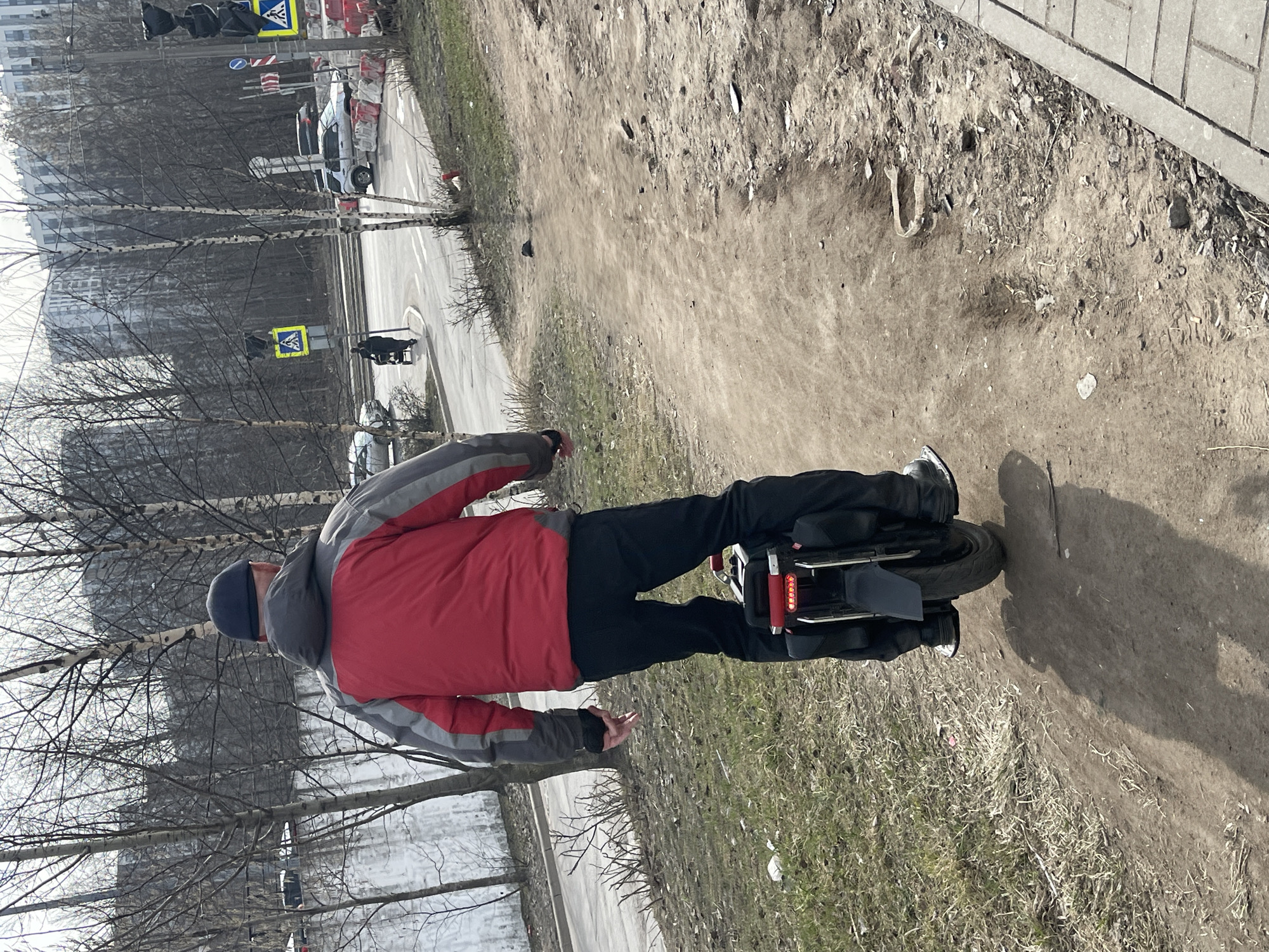 A man is riding an electric unicycle on a dirt path in Saint Petersburg, Russia. He is wearing a red and grey jacket, black pants, and a dark blue beanie. The path is alongside a paved sidewalk and a road with traffic lights and pedestrian crossings visible in the background. The trees are bare, suggesting it is late autumn or early spring. In the distance, there is a building under construction surrounded by traffic barriers.