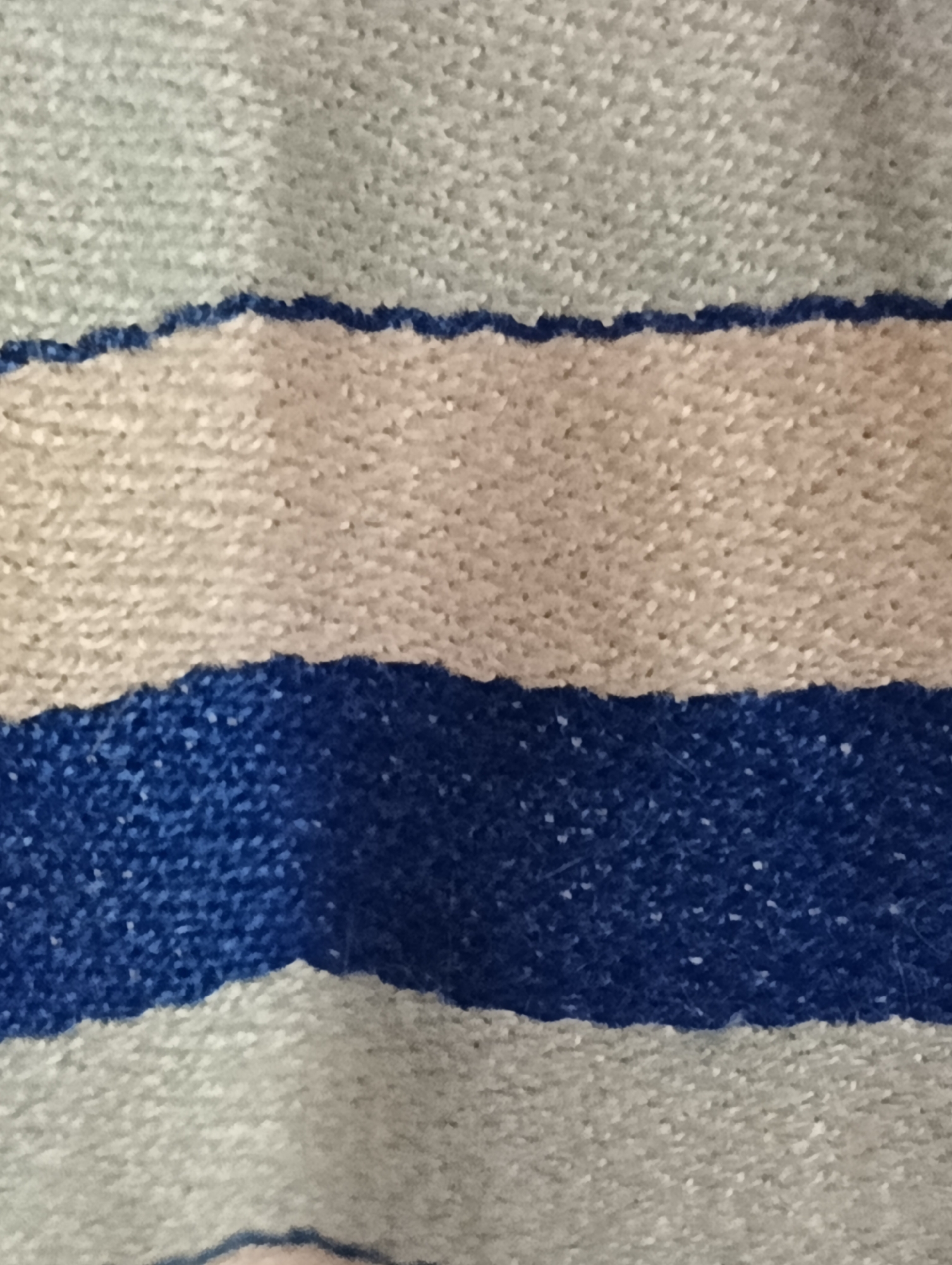 The image presents a close-up view of a textile or fabric featuring horizontal stripes. The dominant colors are light gray, beige (or light tan), and a vibrant royal blue. The top and bottom sections of the image show the light gray color, while a central beige stripe is visible, bordered above and below by blue stripes.
The texture of the fabric appears soft, possibly knitted or woven, with visible individual fibers and a slightly napped or fuzzy quality, particularly noticeable along the edges of the color transitions. These edges are not perfectly straight but show a slightly irregular, soft, or organic ripple, characteristic of textile construction. Small white flecks are dispersed across the surface of all colors, especially prominent in the blue stripe, which could be lint or part of the yarn composition.
There are no people, identifiable objects other than the fabric, or any discernible activity taking place. No text is visible anywhere in the image. The lighting is even, without harsh shadows, suggesting an indoor setting or soft natural light. The provided location context of Biliaivka, Ukraine, cannot be confirmed or inferred from the visual information within the image itself.