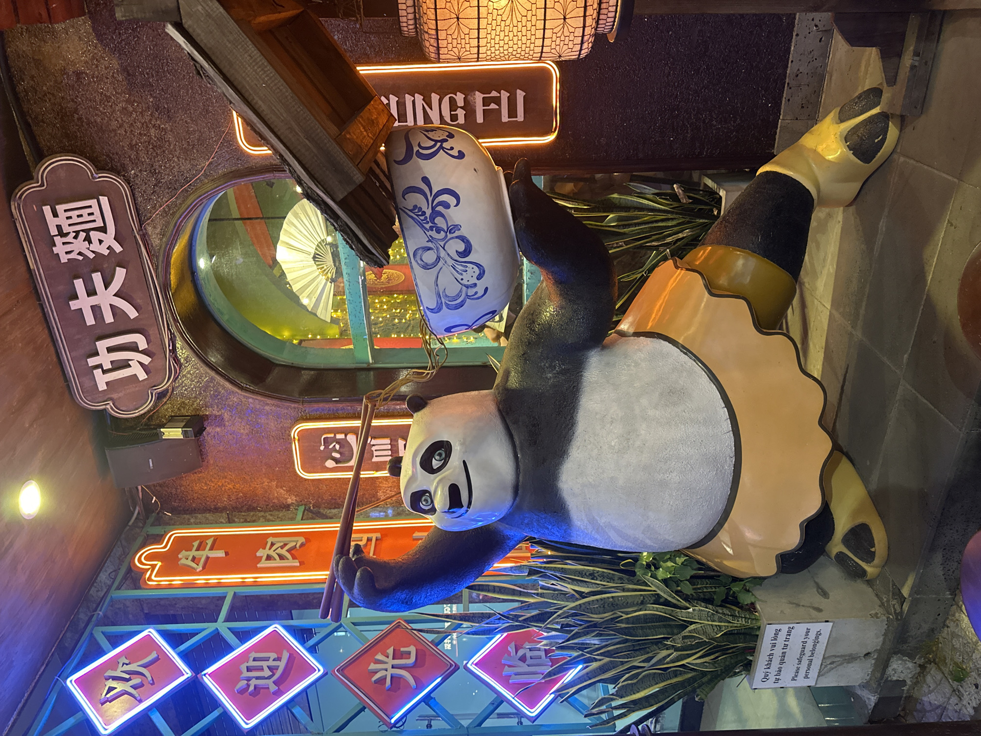This image depicts a large statue of Po from Kung Fu Panda holding chopsticks and a bowl of noodles. The statue is positioned in front of a building with Chinese-style signage, including "Kung Fu Noodles" in Chinese characters above an arched window. Several neon signs with Chinese characters are visible on the left, including "Welcome" and "Beef Noodles." The statue is adorned with a tan apron and yellow footwear. In the foreground, there is a plant in a concrete pot with a sign in Vietnamese and English that reads "Please safeguard your personal belongings." The overall setting suggests a restaurant or themed establishment in Da Nang, Vietnam, with a distinct Asian cultural influence.