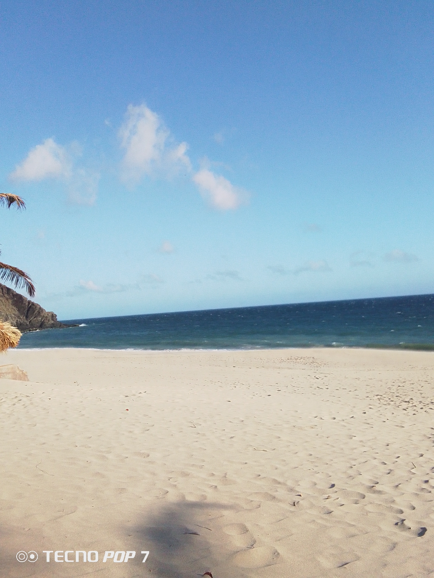 The image depicts a beach scene in Pedro González, Venezuela. The foreground consists of a wide expanse of light-colored sand marked with various footprints and shadows. The sand appears dry and undisturbed. In the midground, the dark blue sea meets the sand with small, gentle waves. In the background, a rocky outcrop juts out into the sea. Partially visible on the left side of the image are some palm leaves and a thatched roof structure. The sky is a clear, light blue with scattered small, white clouds. The overall impression is of a serene, deserted beach on a sunny day. A "Tecno Pop 7" watermark is visible in the lower-left corner.