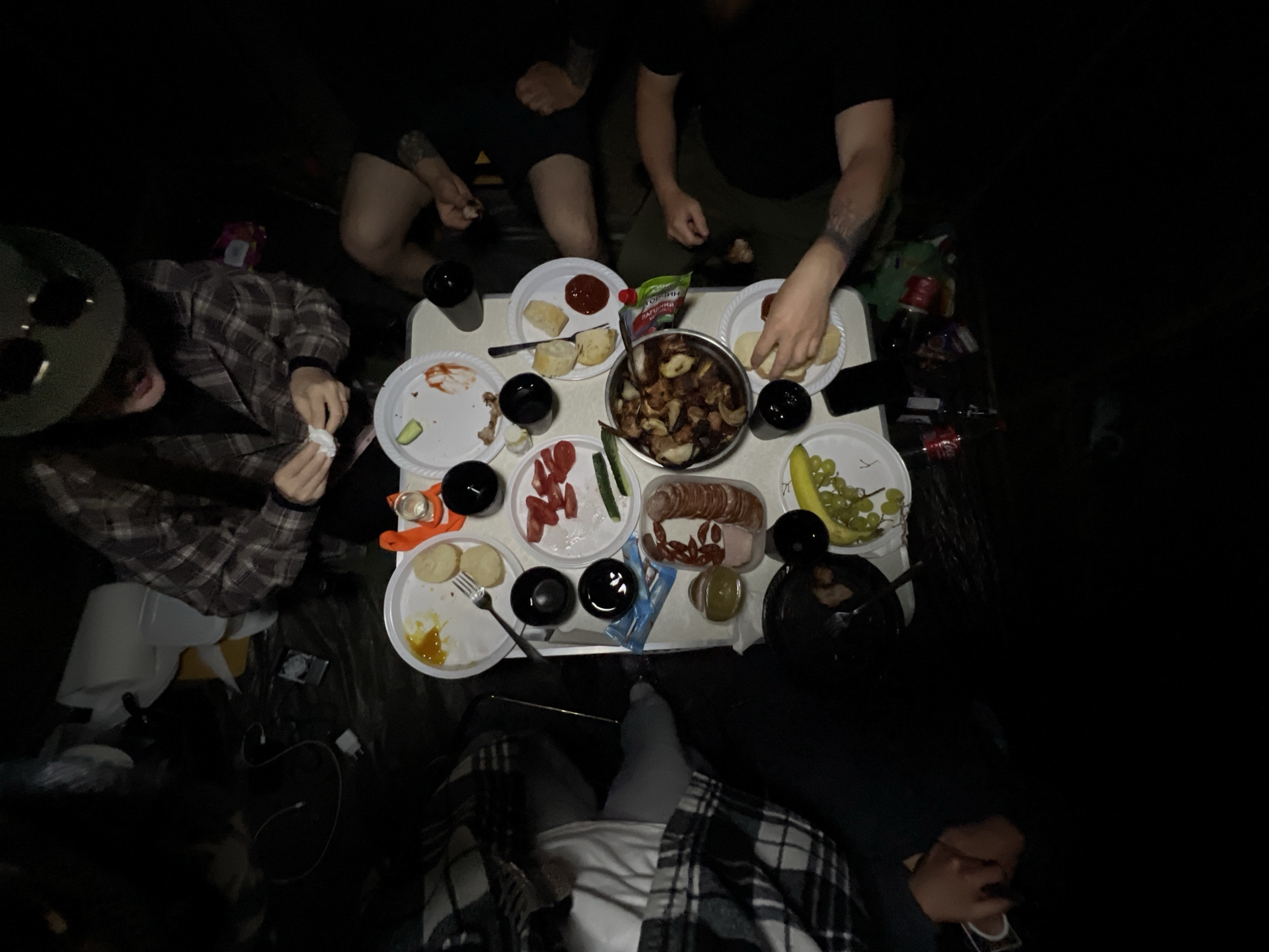 The image shows an overhead view of a group of people sitting around a table laden with food. The scene appears to be indoors, possibly in a dark or dimly lit area. There is no information about the location in Romanove Selo, Ukraine in the image.
The table holds multiple plates of food, including sliced bread, tomatoes, cucumbers, sausage, potatoes, and a bunch of grapes. There are also several black cups or glasses scattered around the table, and what appears to be a bottle of ketchup.
Around the table, several people are visible. Some are reaching for food on the table, while one person on the left is holding a napkin. The attire of the people varies, with some wearing t-shirts and others in flannel shirts.