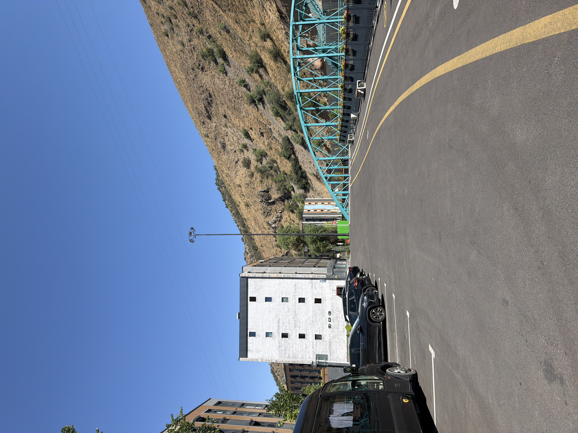 A wide, clear blue sky stretches above a dry, brown mountainside dotted with sparse green vegetation. In the foreground, a paved road with yellow and white lane markings curves to the right. To the left, a row of parked black cars, including a van, are situated in marked parking spaces. Behind the cars, a modern, white building with a dark roof and numerous square windows stands tall. Further to the left, the facade of another modern building, with multi-colored vertical stripes, is partially visible. In the distance, a vibrant teal-colored pedestrian bridge with intricate latticework spans across what appears to be a dry riverbed or ravine, with planters lining its sides. A tall lamppost stands near the center of the scene. The overall environment suggests an urban area integrated with natural mountainous terrain in Khumsan, Uzbekistan.