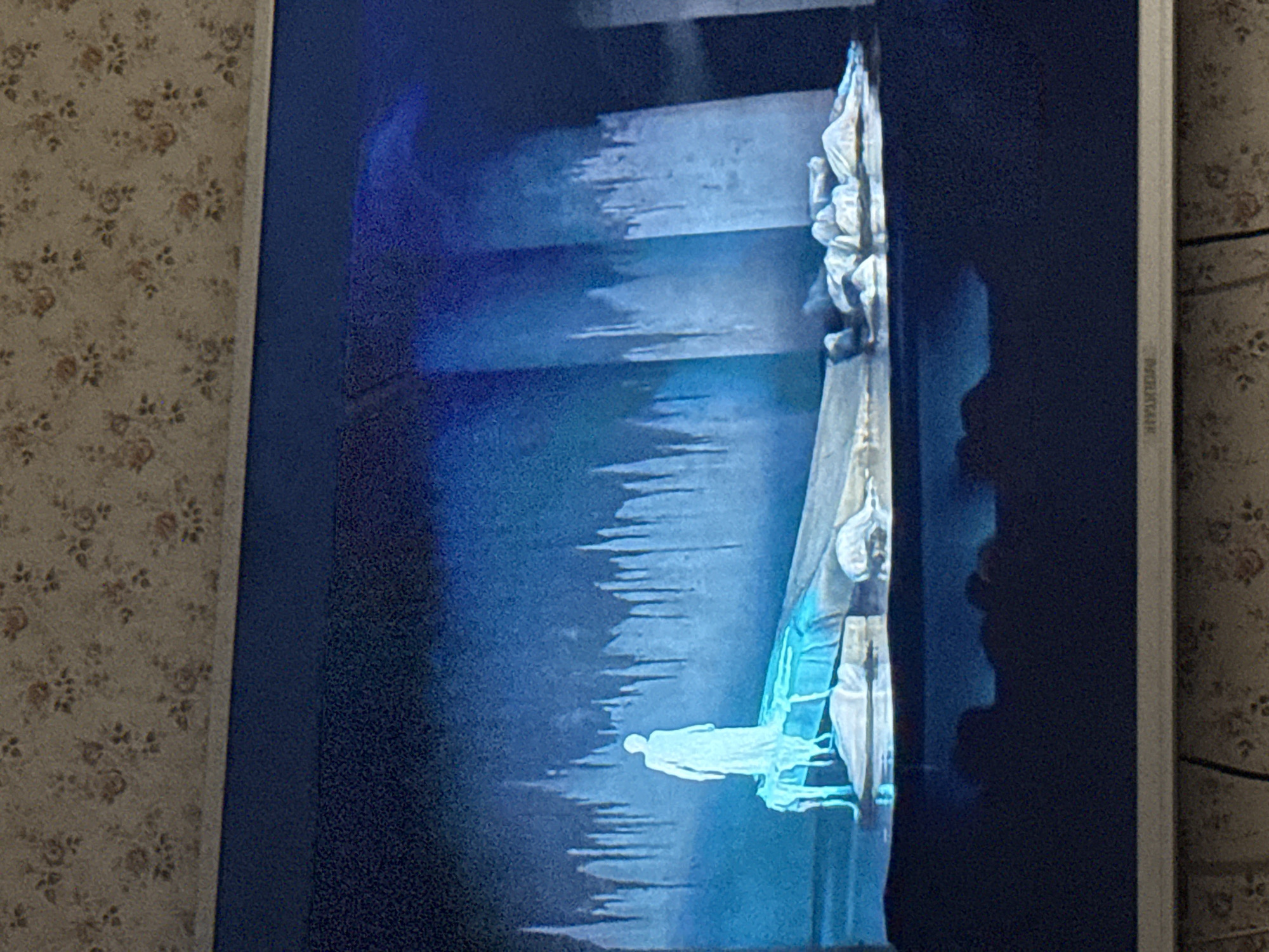 A television screen displays a scene from what appears to be an opera or theatrical performance. The scene is dominated by blue and white hues. Central to the view is a spectral, glowing figure of a person lying horizontally, seemingly floating in the air. Above this figure are several horizontal, step-like levels that create a sense of vertical depth. There are other figures in white costumes positioned on the right side of the scene, close to what might be a wall or a backdrop. The scene is shadowed on the right, possibly indicating lighting effects or the presence of an object casting a shadow. The image itself is a photograph of a TV screen, with a floral patterned wallpaper visible in the background on the left, and the television's border on the right. The location of Mytishchi, Russia, is not discernible from the details present in the image.