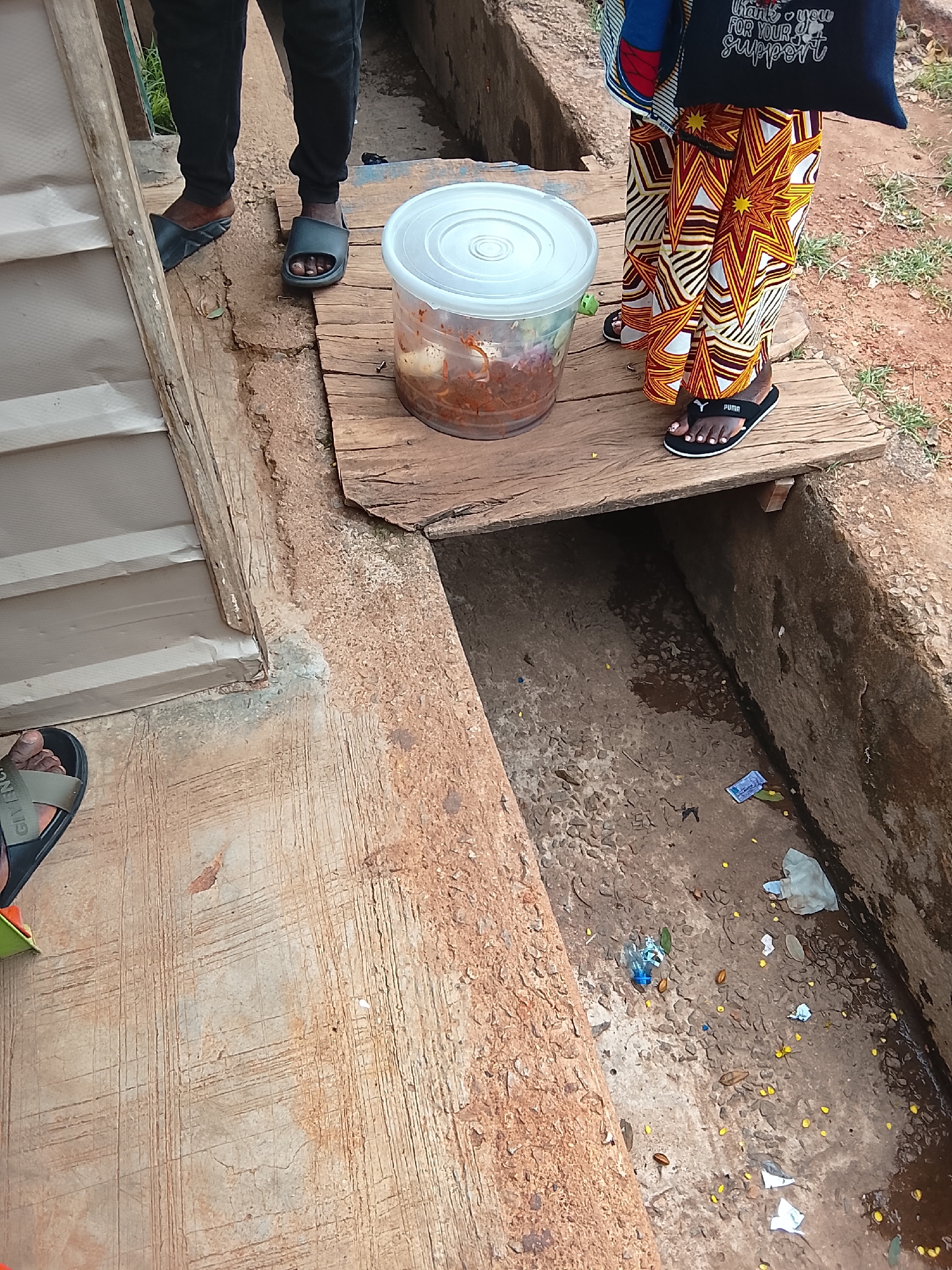 In Jos, Nigeria, two individuals stand on a wooden platform overlooking a drainage ditch. The person on the left wears black trousers and dark slides. The person on the right wears a colorful patterned skirt and black Puma slides. In front of them, a clear plastic container holds food, possibly eggs and a red sauce, resting on the wooden planks. The drainage ditch below is filled with debris, including plastic wrappers, bottle caps, and fallen yellow flowers, indicating an outdoor urban environment.