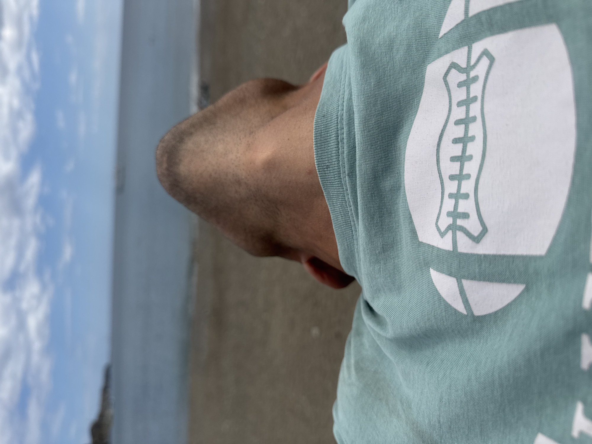 This is a close-up, low-angle shot of a person's neck and the back of their head, wearing a light blue t-shirt with a white football graphic. The person is looking upwards, with their head tilted back. The background is out of focus, showing a beach, the sea, and a cloudy sky. The scene is likely outdoors. The location is Port-Vendres, France.