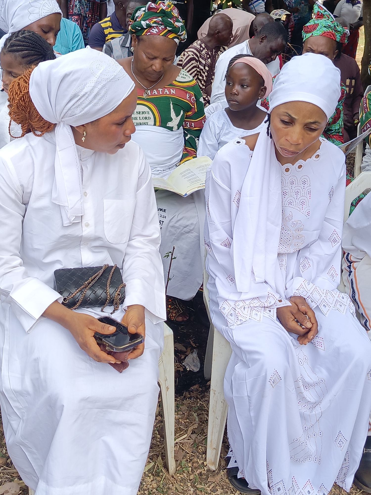 This medium shot captures an outdoor gathering, likely a religious service or community event, in Andaha, Nigeria. Two women in white robes and head coverings are seated prominently in the foreground. The woman on the left wears a plain white long-sleeved garment and a white headscarf, with visible light brown braided hair and gold hoop earrings. She holds a smartphone, resting her hands on a dark handbag with a gold chain strap. She appears to be conversing with or looking at the woman on her right. This second woman is dressed in an ornate white long-sleeved dress featuring intricate cut-out patterns on the sleeves, neckline, and lower portion, with a white headscarf draped over her shoulder. Her gaze is directed downwards, suggesting contemplation or listening.
Both women are seated on white plastic chairs on dry, earthy ground with scattered leaves. In the background, numerous other individuals, including adults and a child, are visible. Many attendees wear vibrant traditional African attire and head wraps. A young girl in a pink headband and white dress sits between the two main subjects. Several people are holding open books or documents, one clearly displaying "LUKE 3:24" and "CHRIST," indicating religious material. Other discernible text on patterned clothing includes "MATAN", "E SERVE", "HE LORD", "TAWA", "EC", "FE", and "CWA". The scene is well-lit by natural light, suggesting it is daytime, and the overall mood appears calm and attentive.