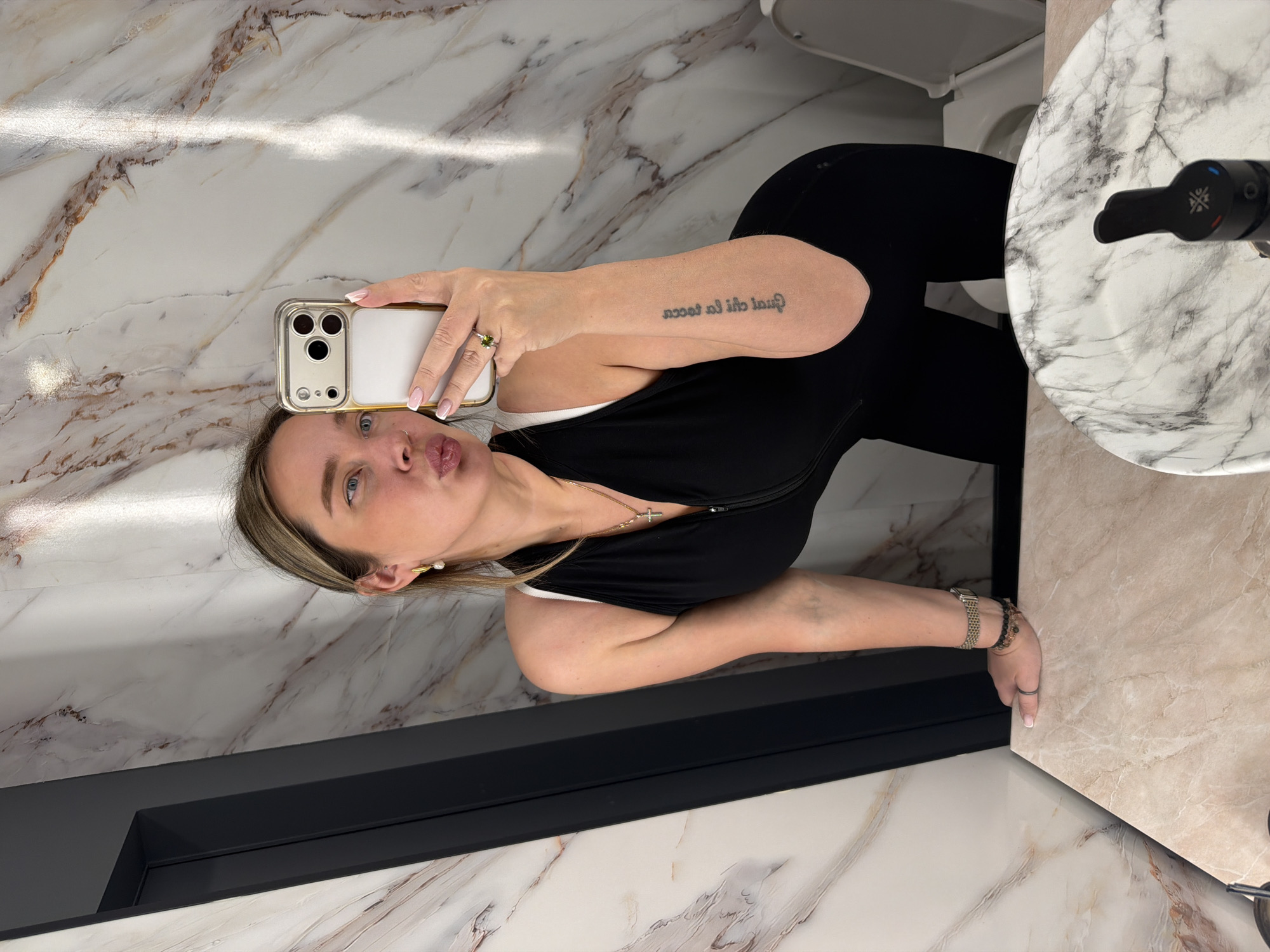 A woman is taking a selfie in a bathroom. The bathroom has a white marble countertop and walls, with a black rectangular fixture below the wall. The woman is wearing a black top, a silver necklace with a cross pendant, and a watch and bracelets on her right wrist. She is holding a smartphone with a gold-colored case. The location is not identifiable as Moscow, Russia, based on the image.