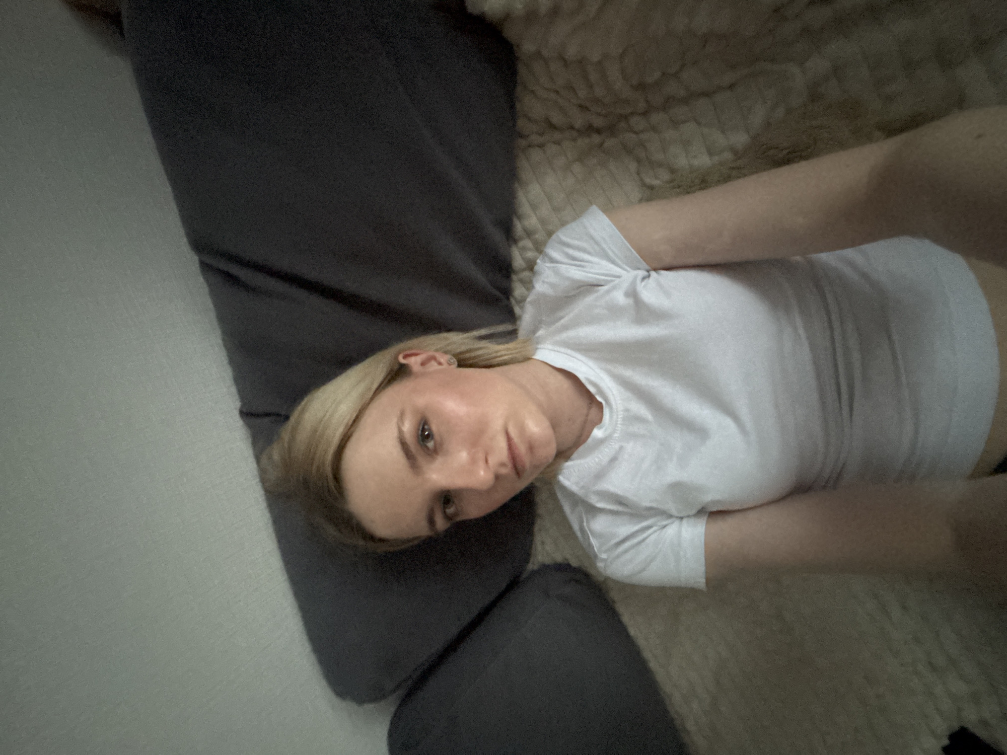 A person is lying on a couch. The couch has gray cushions and a beige textured blanket. The person has blond hair and is wearing a white t-shirt. The location cannot be determined as the scene is indoors and no identifying elements are present to confirm the city of Vsevolozhsk, Russia.