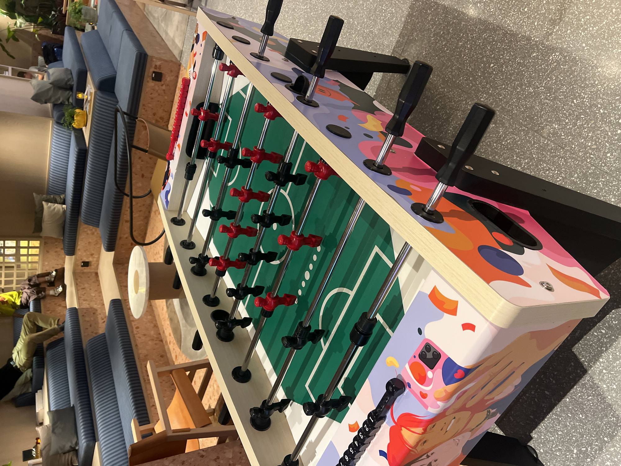 The image captures a foosball table with red and black players in a recreational area. The table features a colorful, artistic design on its sides. In the background, there are tiered blue and grey striped benches, with people seated on them. An orange armchair is visible to the left. The environment appears to be an indoor lounge or common area, possibly part of a larger complex. The flooring is a speckled grey terrazzo. The image quality is clear, and the lighting suggests an indoor setting. No specific Hong Kong landmarks or indicators are visible in the image.