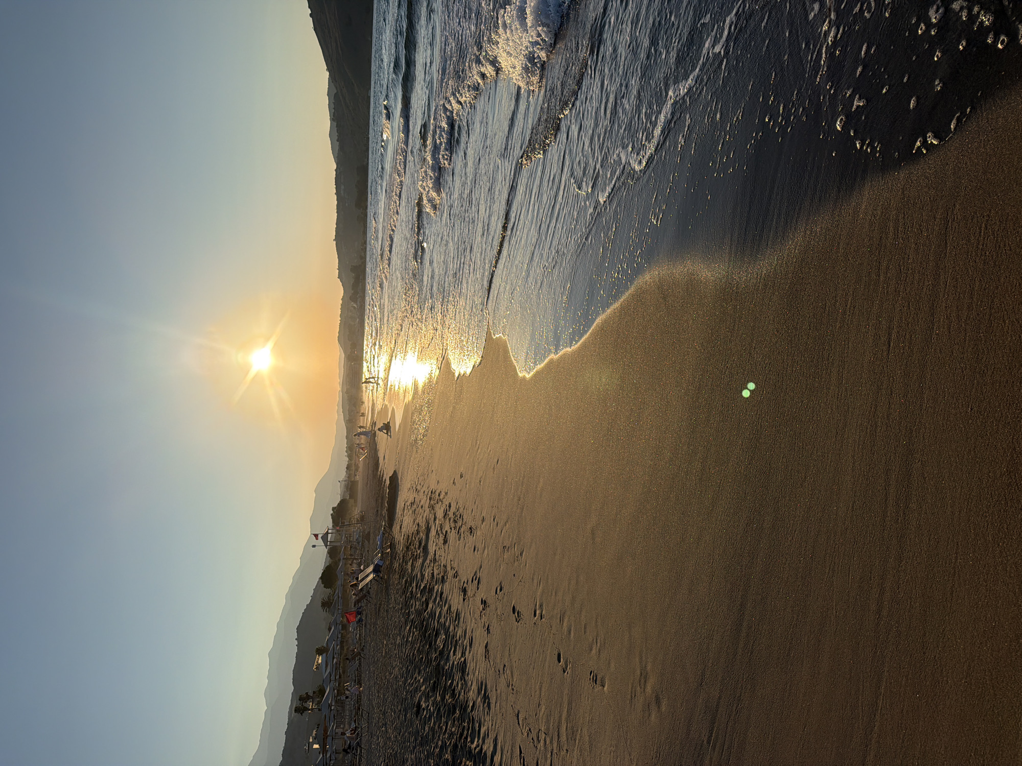 The image depicts a serene beach scene in Georgioupoli, Greece, during sunset. The sun, a bright orb with emanating rays, hangs low in the sky, casting a warm, golden glow across the scene. The sky transitions from a clear, light blue at the top to a soft orange hue near the horizon. Gentle waves roll onto the sandy shore, creating white foam at their crests. Footprints are visible in the damp sand along the water's edge, indicating recent human presence. In the distance, lush green hills and mountains form a scenic backdrop, with some scattered buildings visible along the coastline. A few beach umbrellas and lounge chairs are set up on the sandy expanse, and several figures, presumably beachgoers, are scattered across the sand and near the water, engaged in various activities. The overall atmosphere is peaceful and tranquil, characteristic of a European summer evening.