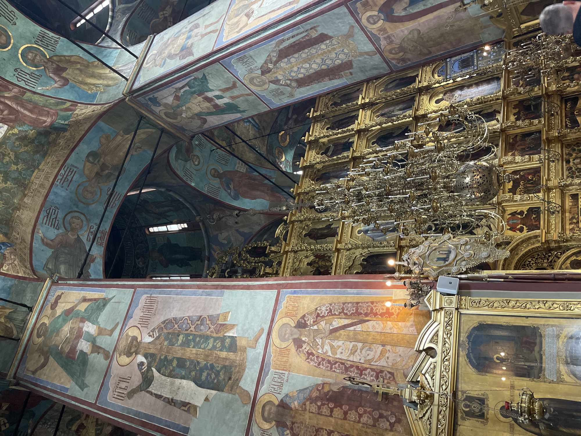 The image depicts the interior of the Dormition Cathedral within the Trinity Lavra of St. Sergius in Sergiyev Posad, Russia. The viewpoint is looking upwards towards the ceiling and a large golden iconostasis. The cathedral walls and ceiling are adorned with vibrant religious frescoes depicting figures in halos, dressed in traditional robes, and carrying scrolls or staffs. The iconostasis, a prominent feature of Orthodox churches, is a richly decorated screen covered in numerous icons, largely gilded, with multiple tiers of religious imagery. A large, ornate brass chandelier hangs centrally, casting a warm glow. Other decorative elements, such as smaller chandeliers and candle holders, are visible. The scene captures the grandeur and spiritual atmosphere of the cathedral, characterized by its religious art and architectural splendor.