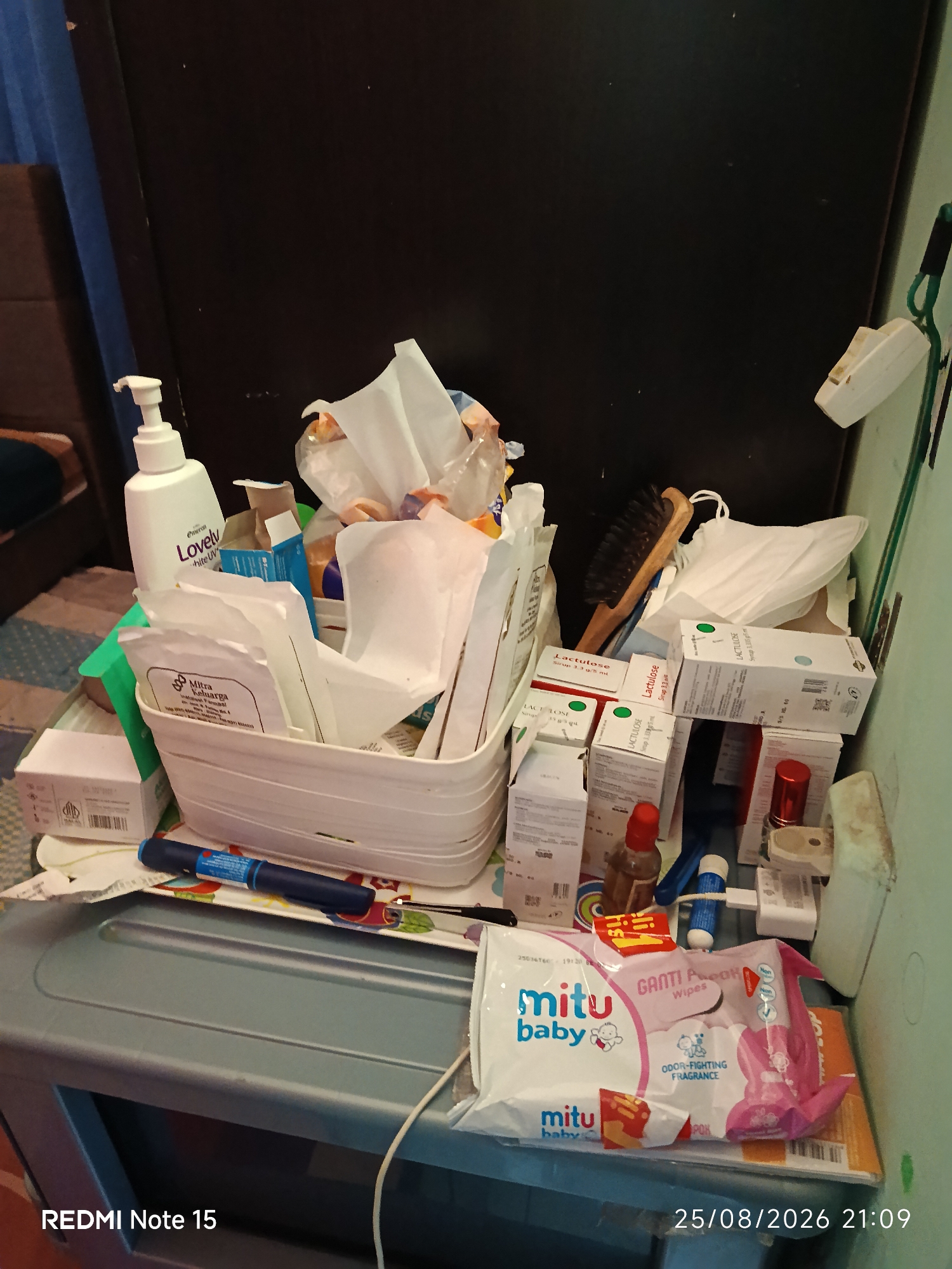 The image depicts a cluttered shelf or surface with various medical and personal care items. Dominating the center is a white plastic basket filled with packaged gauze pads, cotton swabs, and other sterile wound care supplies. To the left, a white pump bottle labeled "Emerson Lovely White UV" is visible, suggesting skincare or lotion. Next to it are several boxes of "Lactulose Syrup" stacked and scattered, indicating medication. Other objects include a small brown bottle, a blue pen-like object (possibly an insulin pen), a toothbrush or hairbrush, and a pack of "mitu baby" wet wipes. The background is a dark, possibly wooden surface. The lower part of the image shows a grey plastic storage container and a digital timestamp reading "25/08/2026 21:09" and "REDMI Note 15". The scene appears to be an indoor setting, likely a bathroom or a personal care area, with a general sense of disarray. There is no information in the image to suggest a specific location within Keboananom, Indonesia.