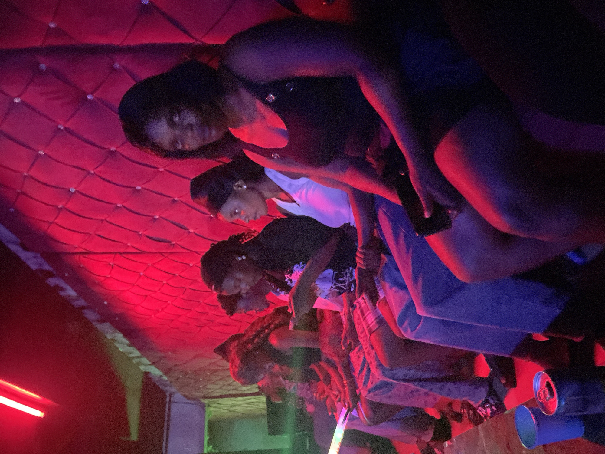 The image depicts a group of individuals, appearing to be women, seated in a dimly lit room with red and purple lighting. The background features a textured, padded wall with diamond-shaped patterns, possibly an entertainment venue or lounge. In the foreground, a blue cup and a soda can are visible on the floor. The individuals are dressed casually, and some are holding or looking at their phones. The overall atmosphere suggests a relaxed or social gathering. The exact location in Durumi, Nigeria is not identifiable from the image alone.