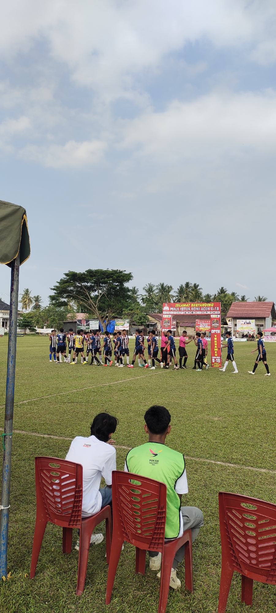 The image shows a soccer field in Kilometer Delapan, Indonesia, with two teams of young players lined up facing each other. A banner in the background reads "SELAMAT BERTANDING PIALA KETUA KONI ACEH U-18" which translates to "GOOD LUCK PLAYING ACEH U-18 CHIEF KONI CUP." Two individuals are seated on red plastic chairs in the foreground, facing away from the camera and towards the field. The sky is partly cloudy.