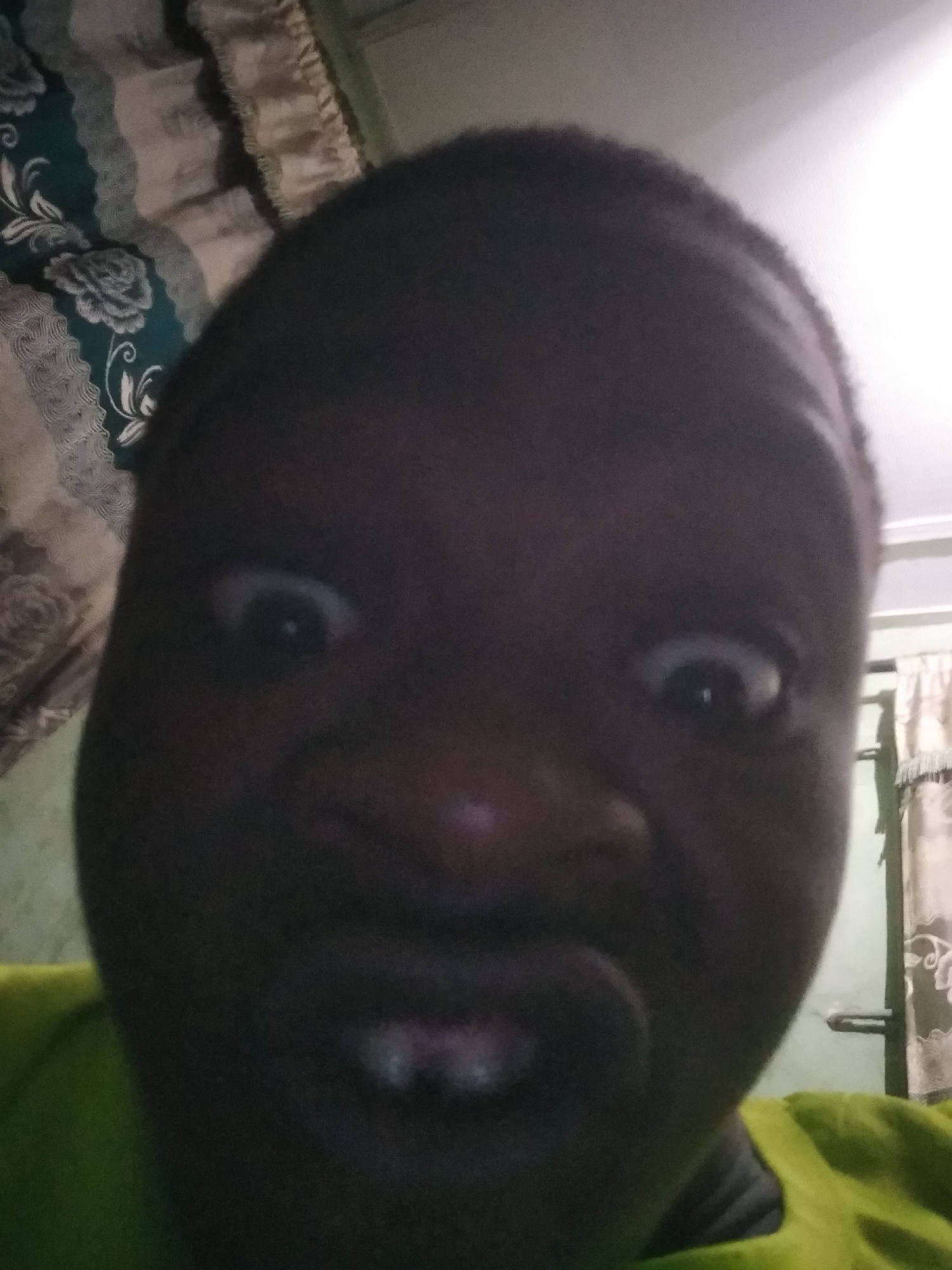 An extreme close-up of a dark-skinned person's face, displaying widened eyes and pursed lips. A green garment is partially visible on the person. A patterned curtain is visible on the left and another behind. The location is Shangisha, Nigeria.