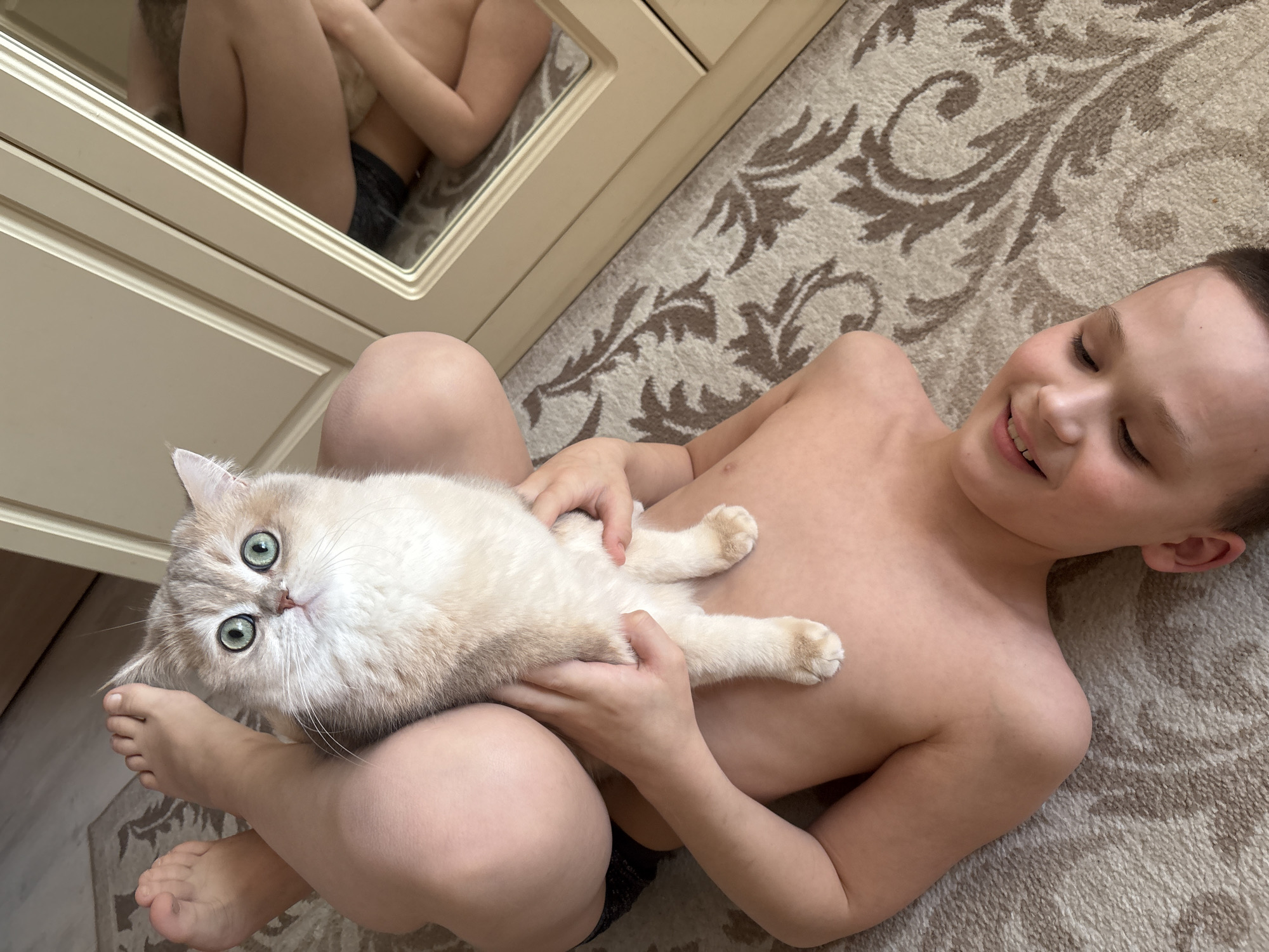 A young boy is lying on a patterned carpet, cradling a light-colored cat in his arms. The boy is fair-skinned, shirtless, and smiling with his eyes closed. The cat has wide, green eyes and is looking directly at the camera. A mirror reflects the boy and the cat from another angle. The location is Chaikovsky, Russia.